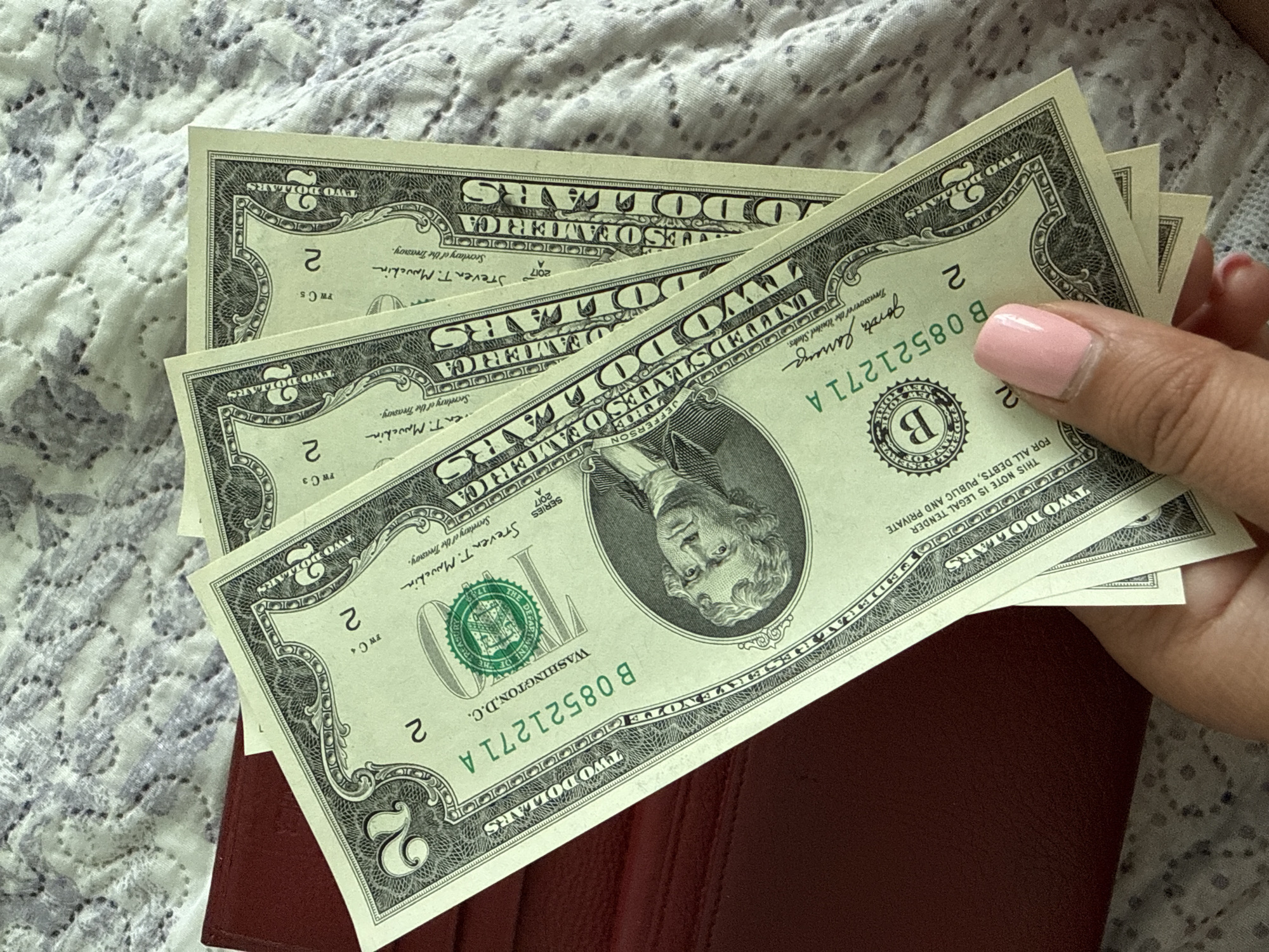 A hand with a light pink fingernail is holding three two-dollar bills, partially fanned out, over a textured, light-colored blanket and a dark red object, possibly a wallet. The bills are oriented diagonally from the bottom left to the top right of the image. The front of the bills, featuring portraits and denominations, is visible. The background is out of focus, with a pattern resembling floral embroidery. The scene is lit from above. The location in Ekzarh Antimovo, Bulgaria, is not determinable from the visual information provided.