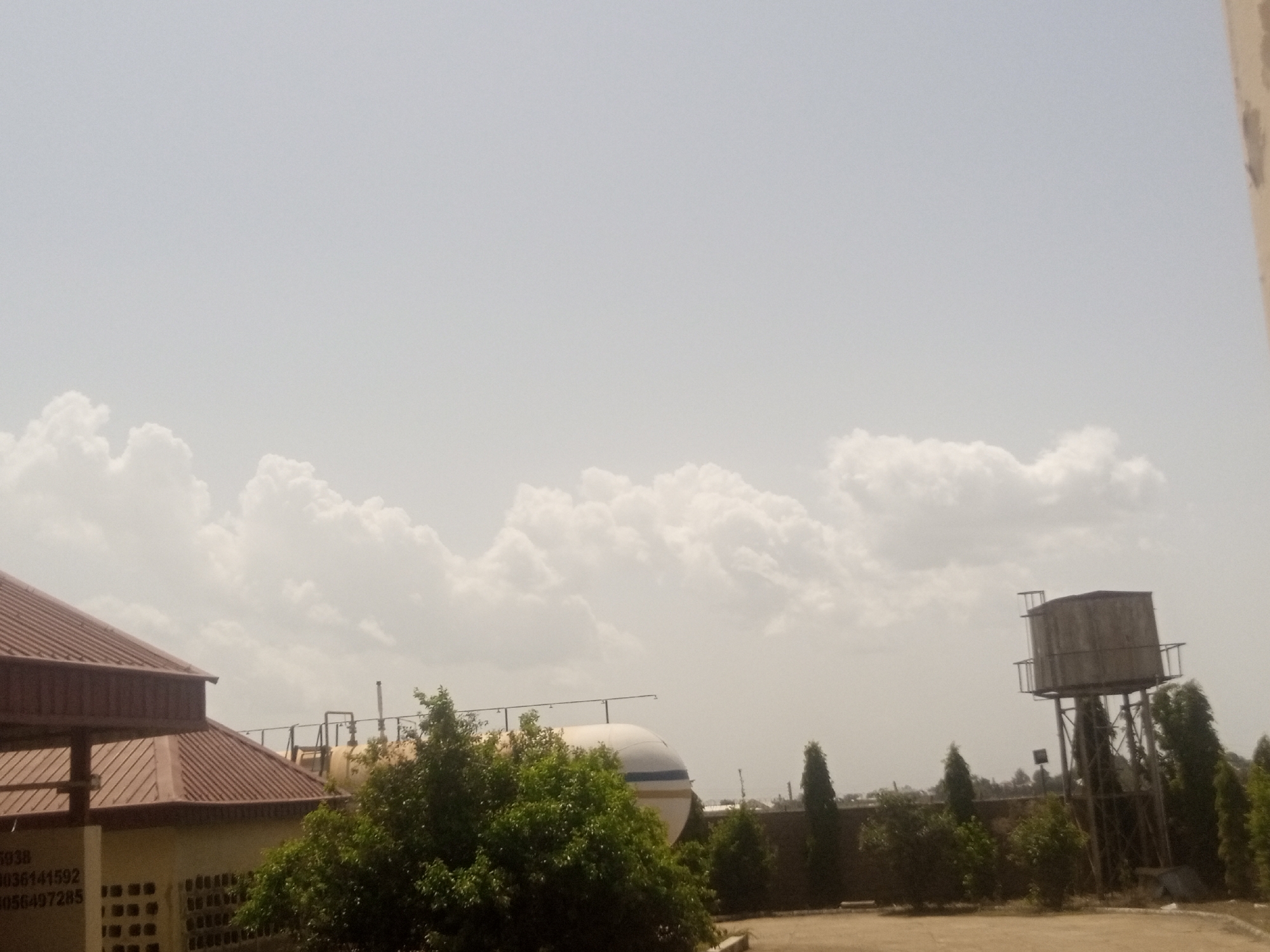 This media file captures an outdoor scene on a cloudy day. The sky is filled with large, puffy white clouds against a pale blue-grey backdrop, suggesting it could be late morning or early afternoon. The setting appears to be an industrial or commercial area, with several structures visible.
In the foreground to the left, a building with a reddish-brown, corrugated metal roof is partially visible. The side of the building is light-colored and has some text inscribed on it. The visible text reads: "3938 036141592 056497285". Adjacent to this building and in front of it, a lush green tree stands prominently, with dense foliage.
Further into the background, a large, white cylindrical object, possibly a tank or silo, is situated. It has a distinctive blue stripe near its top. Above this tank, a network of pipes and supports suggests industrial activity. To the right of the scene, a tall water tower stands, constructed from metal supports and topped with a weathered, square wooden tank. The area around the water tower and behind the tank is dotted with various trees and shrubbery, with some appearing to be tall and slender, while others are more bushy. The ground in the immediate foreground seems to be paved, possibly with asphalt or concrete.
No people or specific activities are depicted in the image. The overall mood is calm and overcast, characteristic of a day with significant cloud cover. The combination of industrial structures and vegetation suggests a location that is functional but also integrated with some greenery.