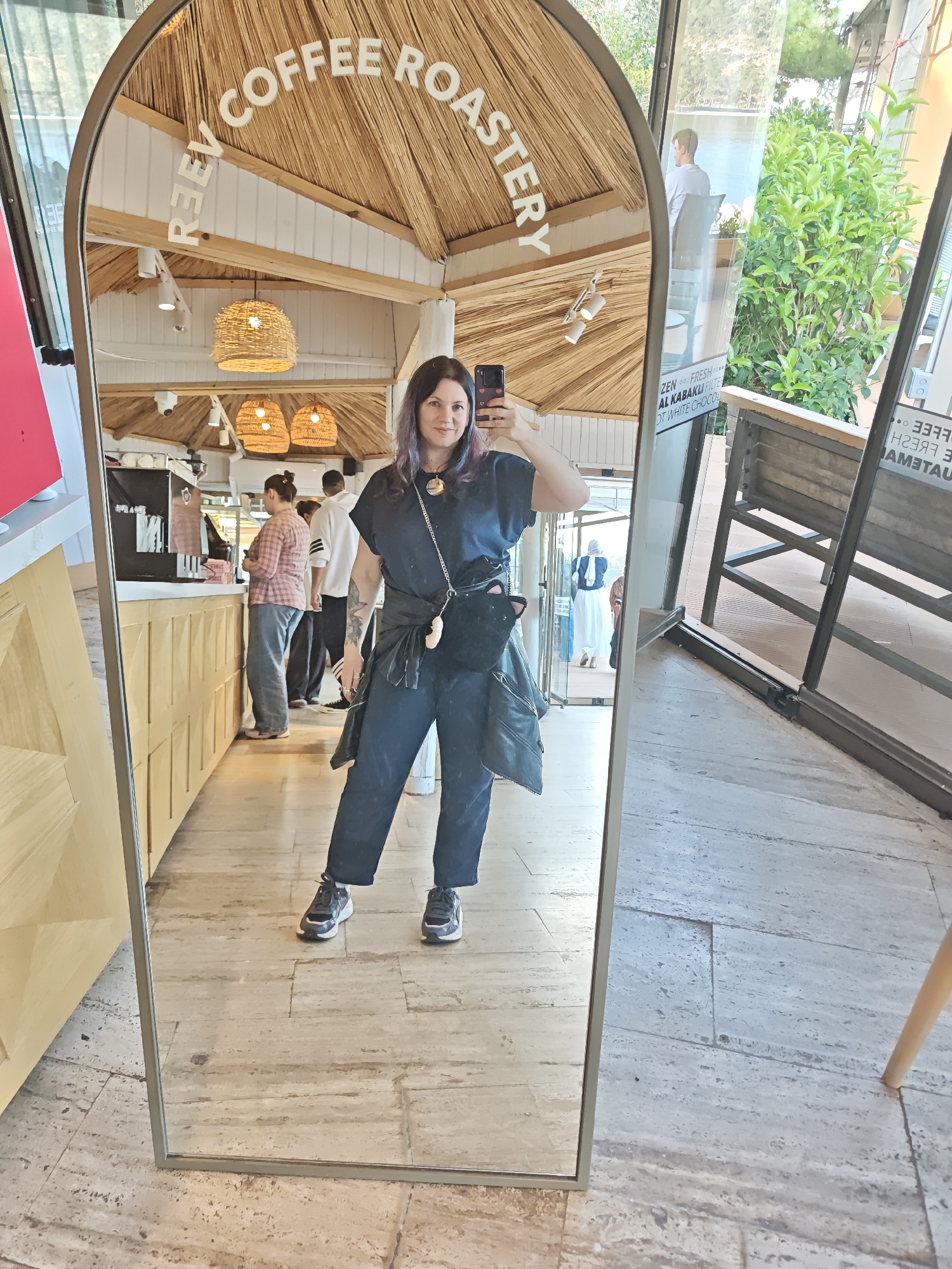 A woman stands in front of a large, arch-topped mirror, taking a selfie in what appears to be a restaurant. The mirror is positioned in front of the establishment, showing the interior of the restaurant with patrons lining up at a counter. The restaurant's ceiling is adorned with thatch, and the words "REV COFFEE ROASTERY" are prominently displayed. Through the glass walls visible in the reflection, lush greenery and an outdoor seating area with a bench are seen. The floor is paved with light-colored tiles. The woman is dressed in a navy blue outfit with a black jacket tied around her waist and black sneakers. The location is likely in the city of Kemer, Turkey, given the context.