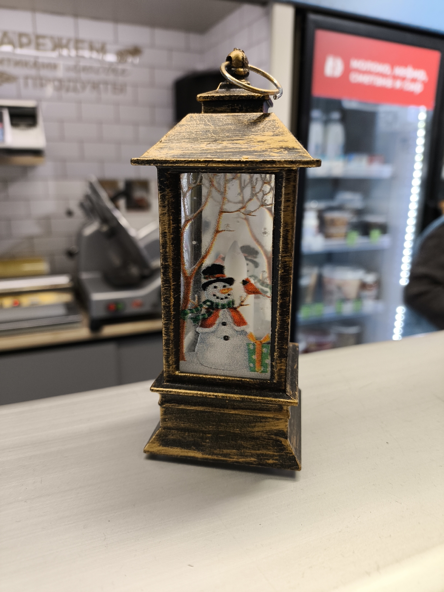 A gold-painted metal Christmas lantern is centered on a light gray countertop. Inside the lantern is a decorative scene of a snowman wearing a black top hat, a green and red scarf, and a red nose, next to a small gift and a small bird. The background depicts bare branches. Behind the lantern is a blurred background with a deli slicer and a refrigerated food display case. A sign in Cyrillic script is mounted on a white tile wall behind the counter. Based on the Cyrillic lettering on the sign, the location is likely in Khimki, Russia.