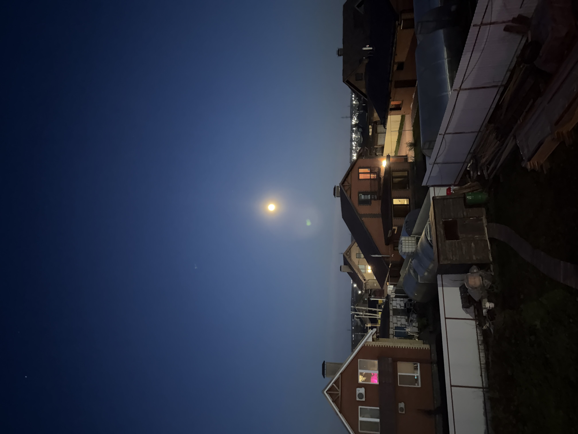 The image captures a quiet night scene in a residential area of Большая Шильна, Russia, viewed from an elevated perspective. The dark blue sky, occupying the majority of the frame, is clear and cloudless, featuring a prominent, bright yellow-white moon slightly to the left of the center. A faint green lens flare is visible near the moon. Along the horizon line on the right, distant lights from a town or city create a soft glow.
Several private houses with sloped, dark roofs are visible in the mid-ground and foreground. Many windows in these houses are illuminated with warm yellow and orange light, suggesting occupied interiors, while one house in the lower part of the frame displays pinkish-purple lights through its windows. In the foreground and mid-ground, various structures can be seen, including sheds, covered areas with translucent corrugated roofing, and stacks of wooden materials, indicating ongoing construction or storage. A large, blue cylindrical tank is also present. Patches of dark green vegetation are visible in the immediate foreground. The scene conveys a sense of peaceful nighttime in a suburban or rural settlement. No people or visible text are present in the image.