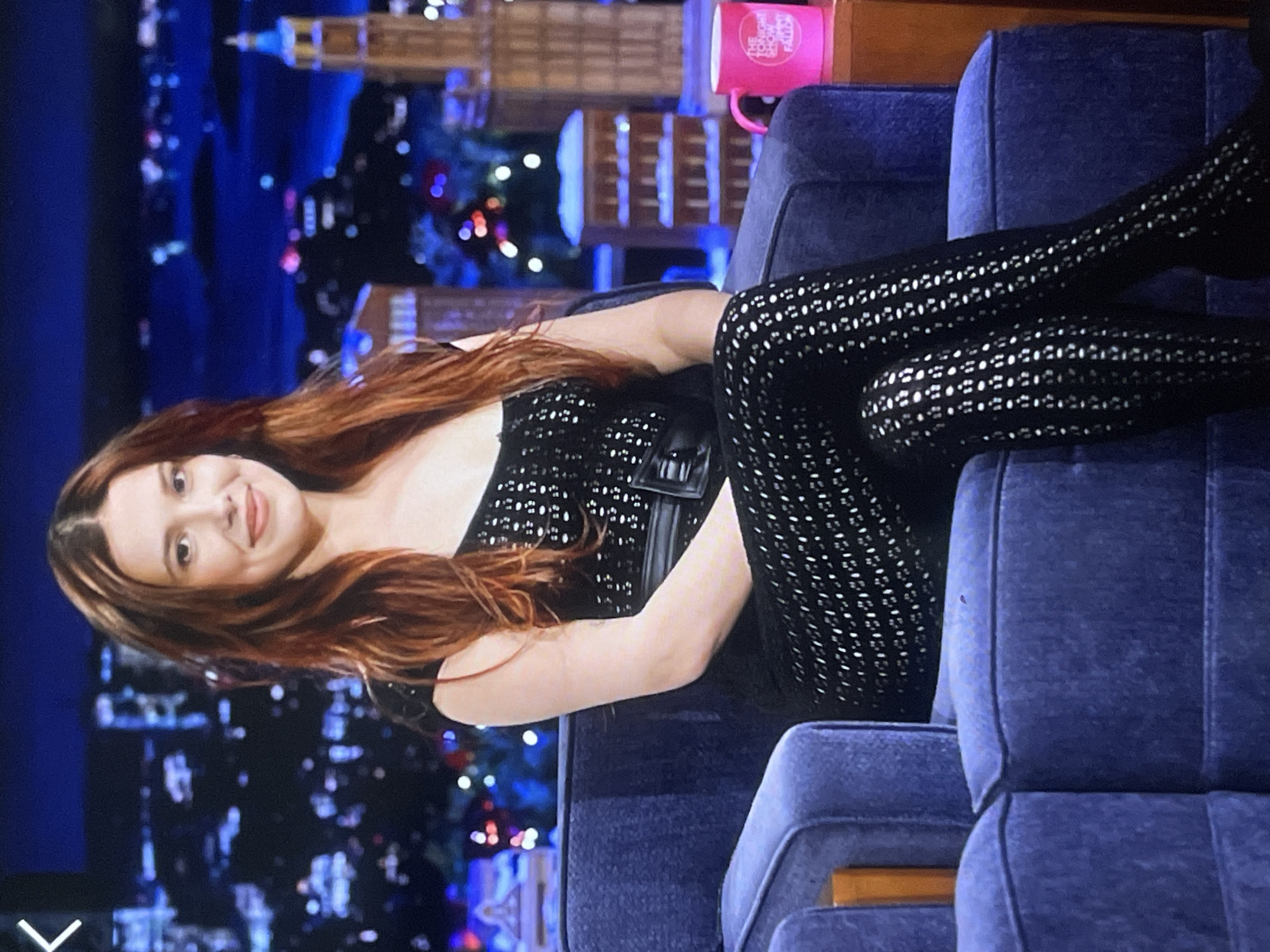 This image captures a young woman seated on what appears to be a television talk show set. The original image is rotated 90 degrees counter-clockwise.
**Subjects:** The main subject is a young woman with long, wavy reddish-brown hair, who is smiling pleasantly and looking towards the viewer.
**Objects:** She is wearing a black outfit, likely a jumpsuit, which features a distinctive pattern of numerous small, light-colored, reflective dots or perforations across its surface. A black belt with a rectangular buckle cinches her waist. She is seated in a plush, dark blue upholstered armchair, with another similar armchair visible next to it. A bright pink mug, bearing white text, rests on a surface behind her.
**Scene and Setting:** The background is a blurred, dark blue cityscape with numerous illuminated windows and structures, creating the impression of a nighttime urban skyline, commonly used as a backdrop for television studios, specifically resembling New York City. The scattered bokeh lights contribute to the nighttime ambiance.
**Activity:** The woman is engaged in what appears to be a television interview or an appearance on a talk show, given the studio setting and her seated posture.
**Notable Details:** The woman's expression is friendly and engaging. The patterned texture of her clothing is a prominent visual detail. The overall lighting suggests a professional studio environment.
**Visible Text:** The pink mug clearly displays the text "THE TONIGHT SHOW STARRING FALLON," identifying the specific television program and its host, which implicitly places the setting in the show's studio, typically in New York City, USA.