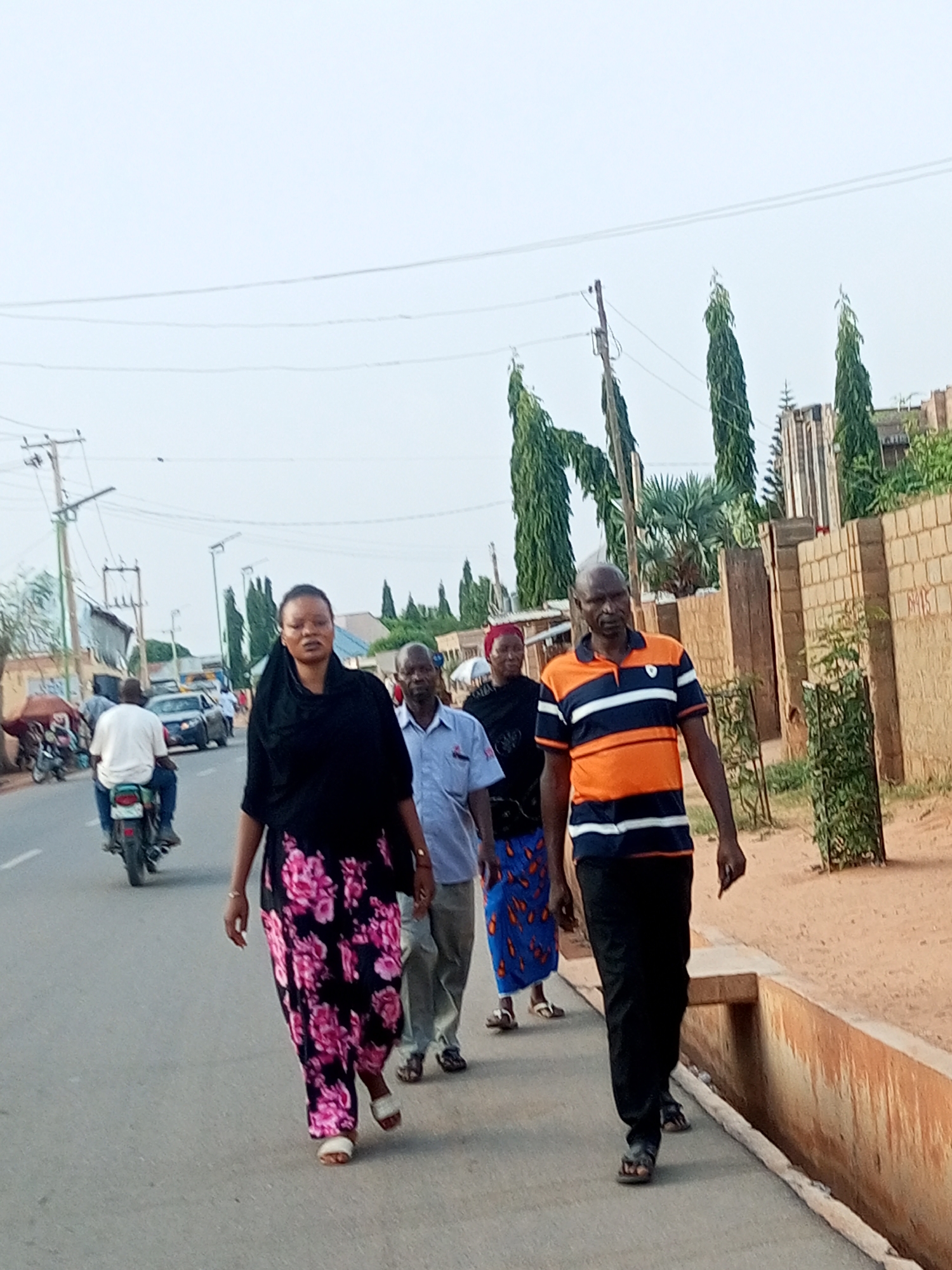 Five people are walking along a road in Tempure, Nigeria. The road has a curb on the right side and traffic on the left. The first person is a woman wearing a black headscarf and a long floral dress. Behind her are two men and another woman. The last person is a man wearing an orange and blue striped shirt and black pants. On the road, a motorcycle is traveling in the opposite direction. Buildings and tall, thin trees are visible along the side of the road. The sky is overcast.