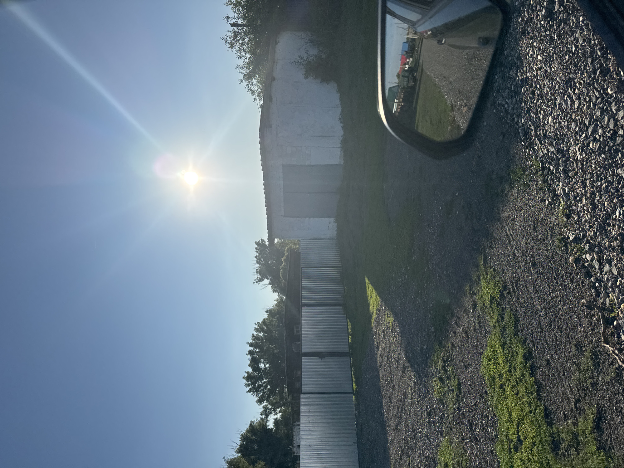 This is a sideways, eye-level outdoor, handheld shot captured on a bright, sunny day. The sun is positioned in the upper left quadrant of the frame, casting a strong lens flare and bright beams across a clear blue sky. In the foreground, the scene is dominated by a gravel driveway. To the right of the driveway, lush green grass grows sparsely between the stones, and a dark shadow is cast by an unseen object.
Dominating the middle ground is a long, low-slung building with a white, slightly weathered facade. In front of the building, and running parallel to it, is a row of what appear to be corrugated metal panels or structures, stacked in a segmented fashion. To the left of this structure, more trees are visible, their green foliage contrasting with the bright sky.
On the far right of the frame, a car's side-view mirror is visible, reflecting a scene of the road ahead with various vehicles and structures. The reflection shows a road, more gravel, and what appear to be colorful buildings in the distance. The overall impression is of an industrial or utilitarian area, possibly on the outskirts of a settlement, given the presence of the gravel, buildings, and what looks like fencing. The time of day is clearly daytime, with the sun high in the sky, indicating it is likely mid-morning to early afternoon. The weather appears to be clear and fair. There is no visible text in the image.