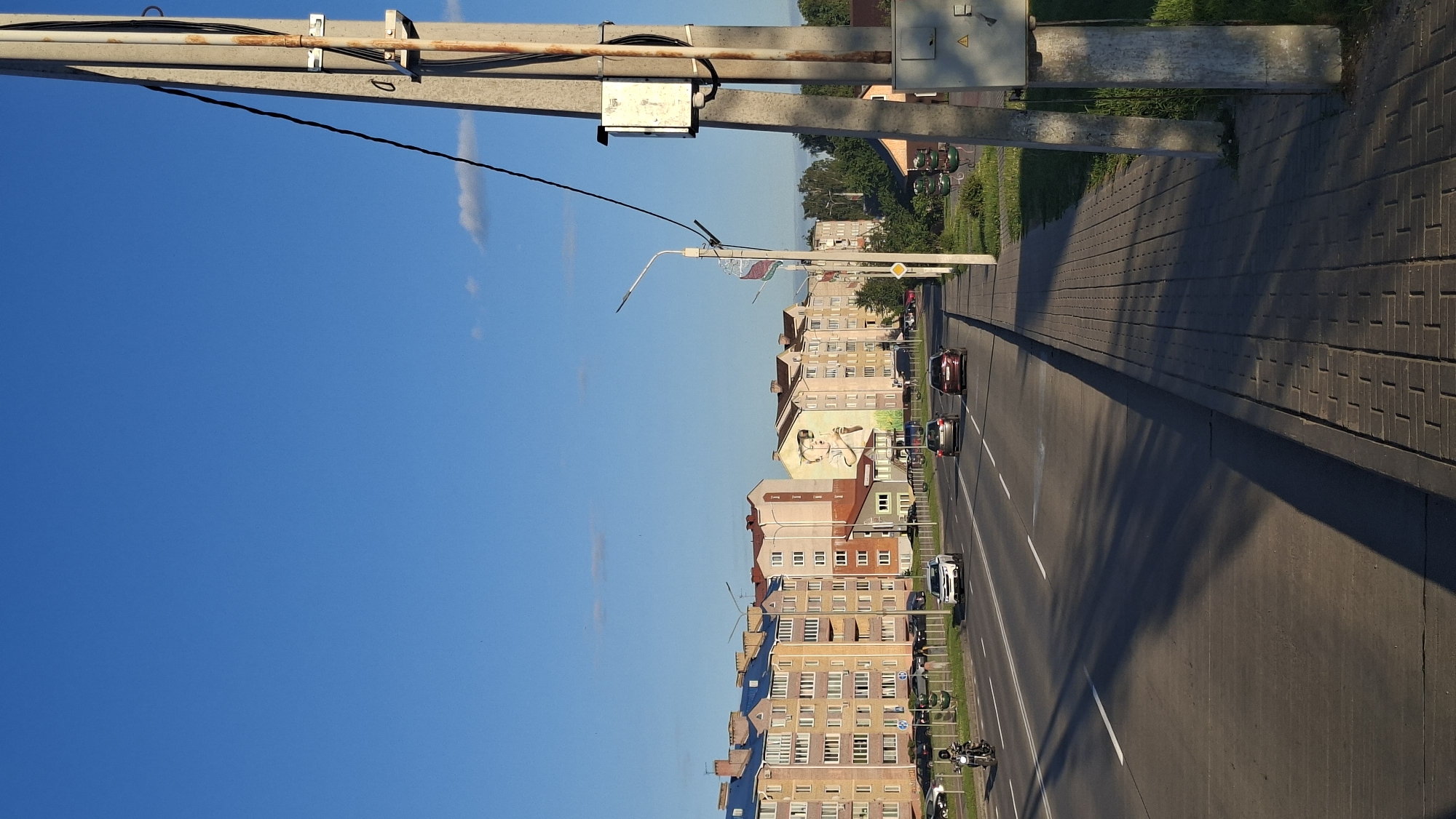 The image depicts a wide, paved road in Smalyavichy, Belarus, with multi-story apartment buildings lining the left side of the road. The buildings are brightly colored with yellow, pink, and blue facades. A mural of a young person is visible on the side of one building. Multiple cars and a motorcycle are driving on the road. On the right side of the road, there is a paved sidewalk, a concrete utility pole with wires, and a green grassy area with trees in the background. The sky is a clear blue with a few wispy clouds. The sun is casting long shadows across the road and sidewalk, indicating it is late afternoon or early evening.