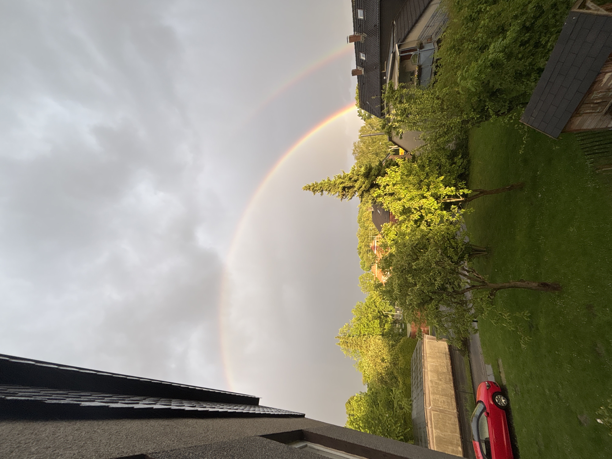 The image shows a residential area in Bochum, Germany, under a cloudy sky with a double rainbow. The perspective is from an elevated position, possibly a balcony or upper floor. In the foreground is a rooftop with dark gray tiles. Below that is a gray facade with a window. To the right, a green lawn is visible, with trees and bushes along a road or pathway. A red car is parked on the grass. In the background, buildings with brown tiled roofs are visible amidst the greenery. The double rainbow arches across the sky from the left side of the frame, terminating behind the trees.