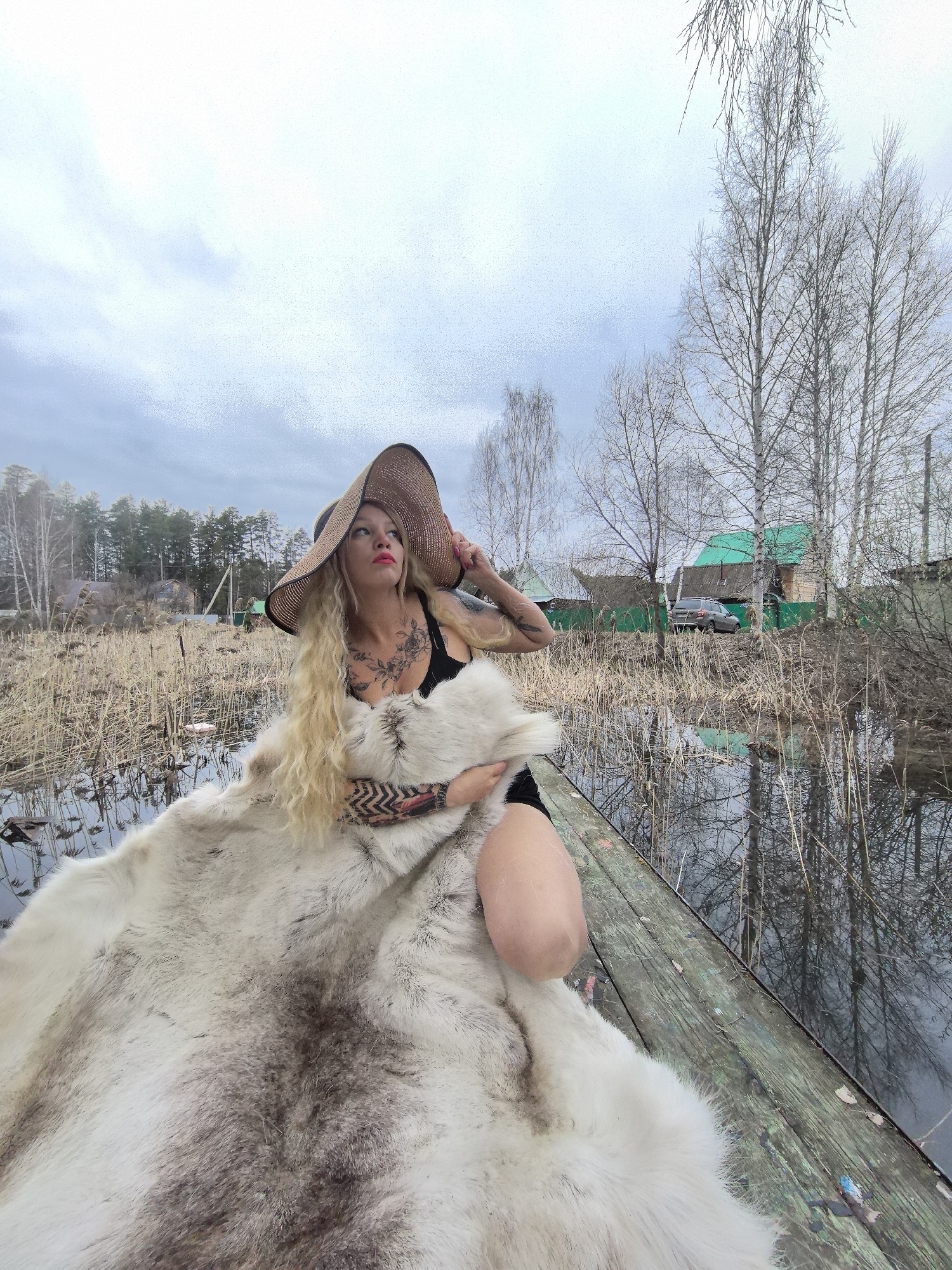 The image depicts a woman seated in a rustic wooden boat on a body of water, likely a flooded area or narrow channel, in Noviy, Russia. She has long, wavy blonde hair and is wearing a black top and a large straw hat, which she holds with her left hand. A large, white animal fur or hide covers her lower body and wraps around her. Visible tattoos adorn her chest, shoulder, and left forearm. Her face is turned upwards and to the left, with bright pink lipstick noticeable.
The scene suggests late winter or early spring, characterized by an overcast, light grey sky and numerous bare deciduous trees, including some that resemble birches, lining the background. The boat is surrounded by dry, tall reeds or grasses growing out of the shallow water. In the distance, several small houses with green roofs are visible, along with a grey car parked near one of them, indicating a rural or semi-rural setting. The water reflects the bare trees. The woman's pose and upward gaze suggest a contemplative moment or a styled photoshoot. There is no visible text in the image.
