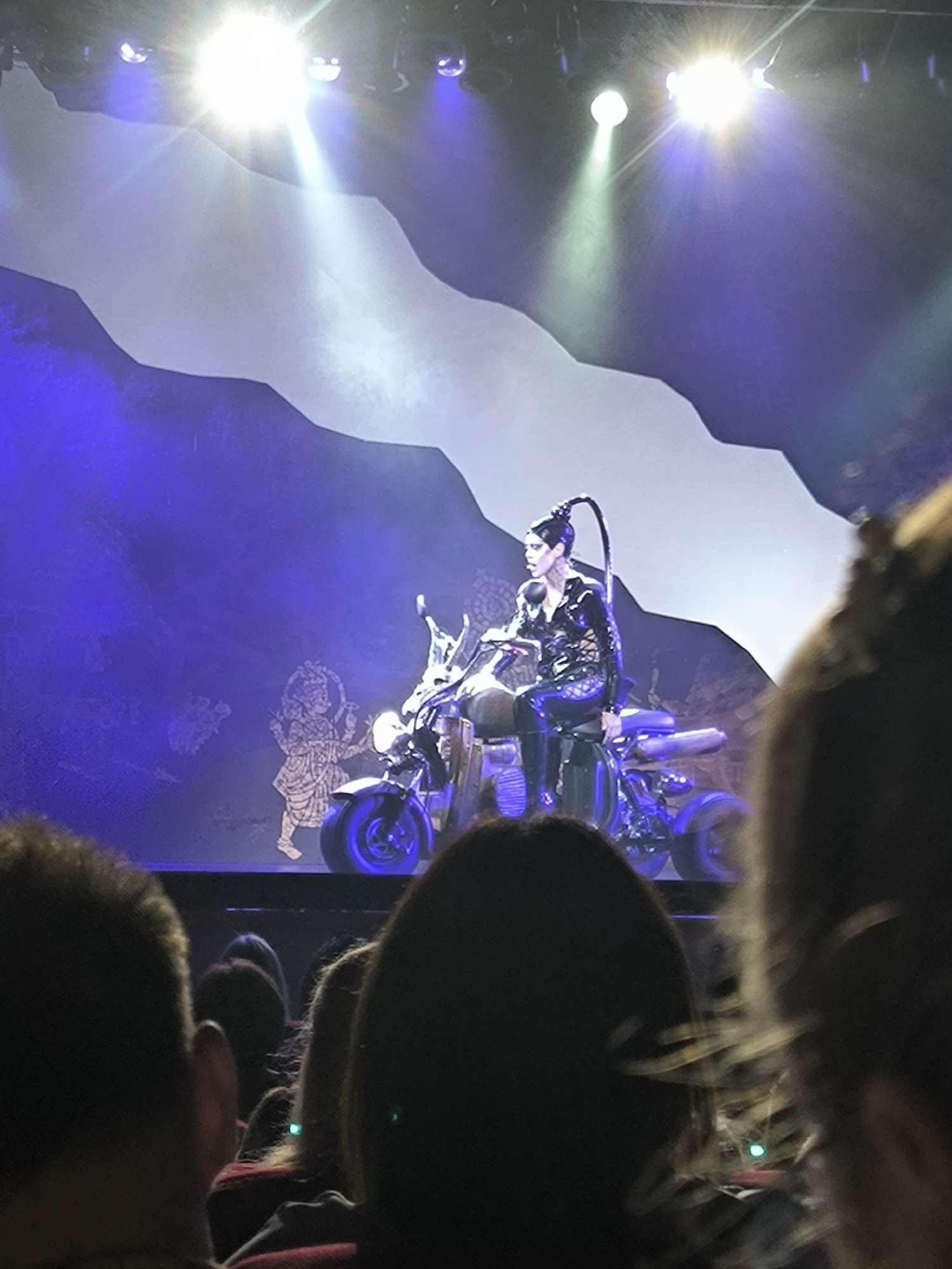 The image depicts a performance on a stage with a dark blue backdrop and stylized white mountain-like structures. A performer in a shiny black outfit, with hair pulled back into a long ponytail, sits on a motorcycle. The motorcycle is dark and has blue accents on its wheels. To the left of the performer, on the backdrop, there are faint images of figures. The stage is illuminated by bright spotlights and purple lighting. In the foreground, the backs of audience members' heads are visible, suggesting the image was taken from within an auditorium or concert hall. There is no visible indication of the specific city or country in the image itself.
