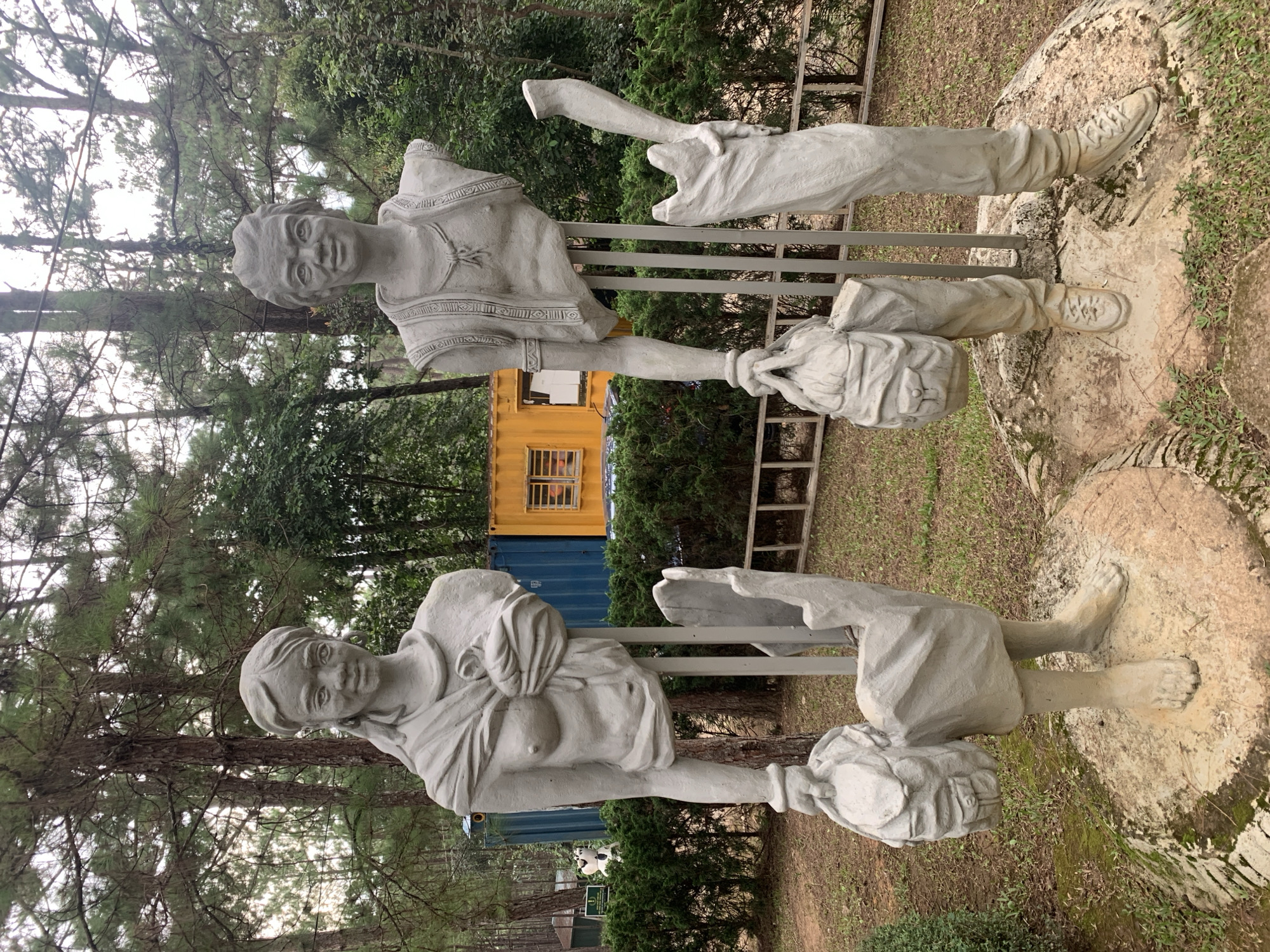 The image shows two concrete sculptures of people in an outdoor setting, likely in Da Lat, Vietnam. The sculptures are displayed vertically, giving the impression of people standing on their heads. Both figures appear to be carrying sacks or bags.
The sculpture in the foreground depicts a person with their torso and head visible. They are wearing a garment that resembles a tunic with decorative bands around the shoulders and chest. Their arms are crossed, and they are holding what appears to be a sack. Their expression is neutral. The lower half of this sculpture, including the legs and feet, is not fully visible.
The sculpture in the background shows the lower body and legs of another person, with their head and upper torso largely obscured. This figure also appears to be carrying a sack. Their legs are bent, and they are wearing what look like boots or laced shoes. The sculptures are supported by metal poles, and are positioned on what appears to be a textured, rough surface, possibly a rock or a constructed base.
Behind the sculptures, there are trees and foliage, suggesting a natural or park-like environment. In the mid-ground, there are shipping containers, one yellow and one blue, with windows visible on the yellow container. A wooden ladder is also visible. The overall scene suggests an art installation or sculpture park. The lighting indicates it is daytime, and the cloudy sky suggests overcast weather. No text is visible in the image.