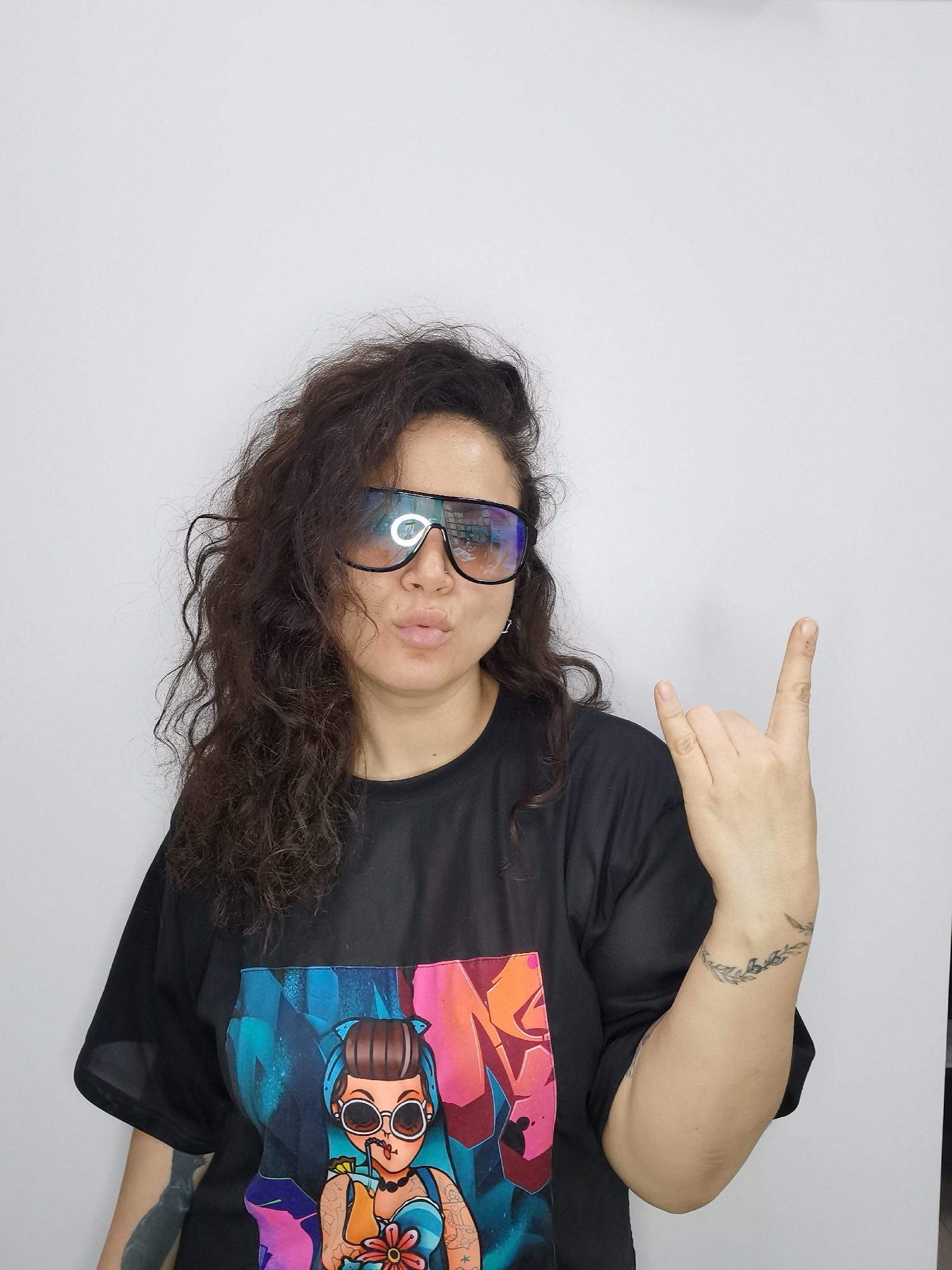 A woman with dark curly hair is posing in front of a white wall, wearing large, mirrored sunglasses and a black t-shirt with a colorful graphic print of a cartoon woman. She is making a "rock on" hand gesture with her right hand and puckering her lips. There is a tattoo visible on her left arm and another tattoo on her right forearm that appears to be a leafy vine design. The lighting in the image is bright, casting minimal shadows. The image is a medium shot, with the woman occupying most of the frame. The background is a plain white wall, with no other objects or discernible features. There is no indication of the location being in Chaikovsky, Russia.