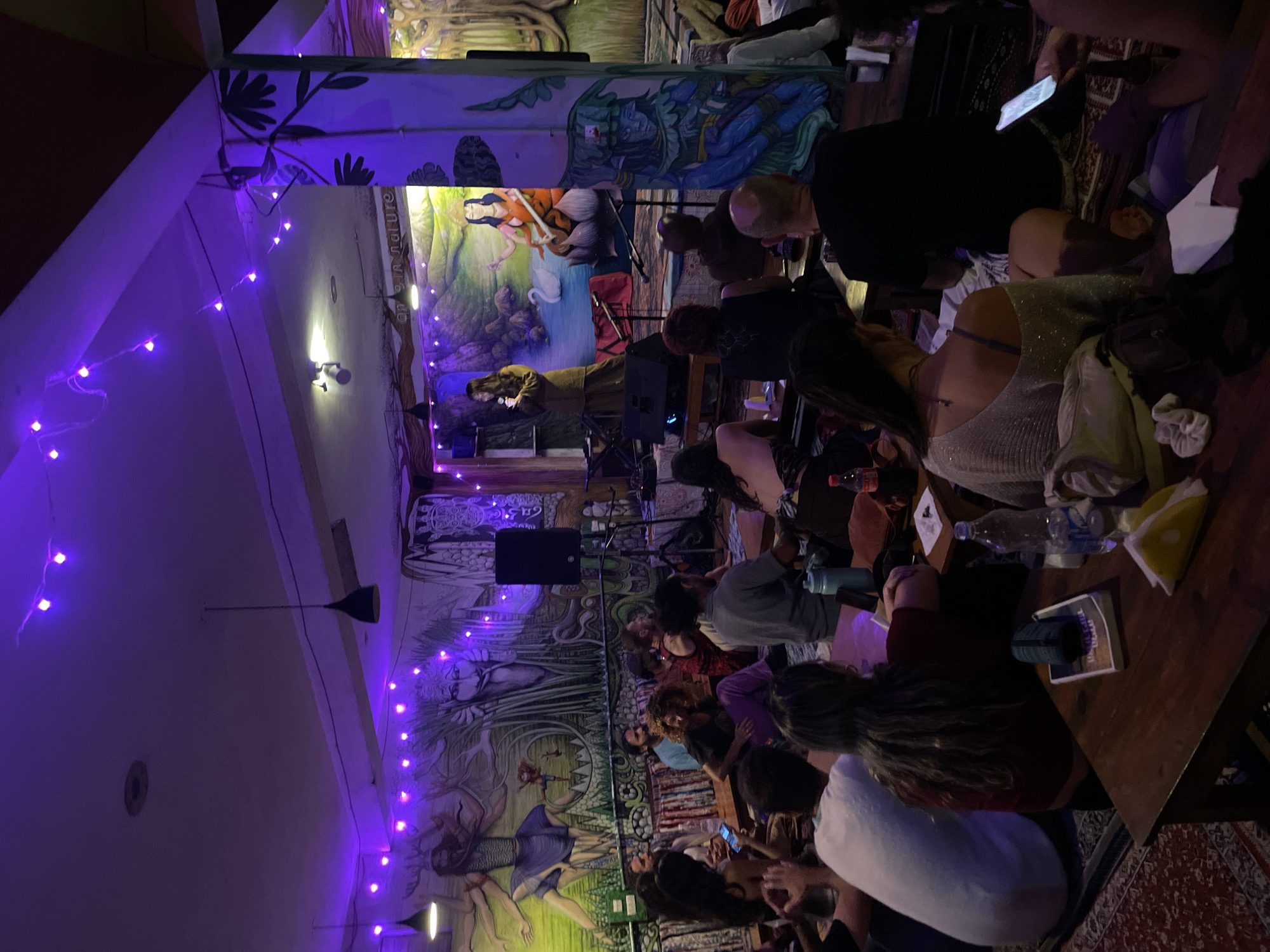 A medium shot captures an indoor event, possibly a performance or presentation, taking place in a dimly lit room adorned with vibrant murals and illuminated by strings of purple fairy lights. The murals, depicting abstract and nature-inspired figures, cover the walls, creating an artistic and bohemian atmosphere. In the foreground, a group of people are seated on the floor, some at low tables, engrossed in the activity on a raised platform at the far end of the room.
On the platform, a woman in a long, flowing brown outfit stands at a podium, appearing to speak or perform. Behind her, a colorful mural features a serene landscape with a woman seated on a lotus flower next to a white swan, a scene suggestive of Indian mythology or spirituality. A keyboard and microphones are also visible on the platform, indicating a musical or spoken-word performance.
The audience consists of individuals of diverse ages and genders, dressed casually. They are seated on cushions and mats arranged on the floor, with some individuals looking towards the platform while others are engaged with their mobile phones. The lighting is primarily from the purple fairy lights, casting a warm and inviting glow over the scene. Several tables, some wooden and others appearing to be cushions on the floor, are scattered throughout the space, holding water bottles, books, and personal belongings. The presence of patterned rugs on the floor further contributes to the relaxed and informal setting. The overall impression is of a community gathering centered around artistic expression or cultural engagement in a uniquely decorated space.