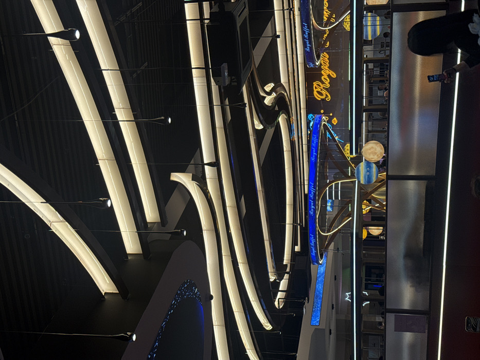 The image shows a modern interior with a dynamic display of linear and curved lights, creating a striking visual pattern against a dark background. A "Royal Buffet" sign is prominently featured, adorned with decorative elements. Reflective surfaces and a glimpse of the restaurant’s interior are visible on the right. People are partially seen, adding a sense of scale and activity to the scene.