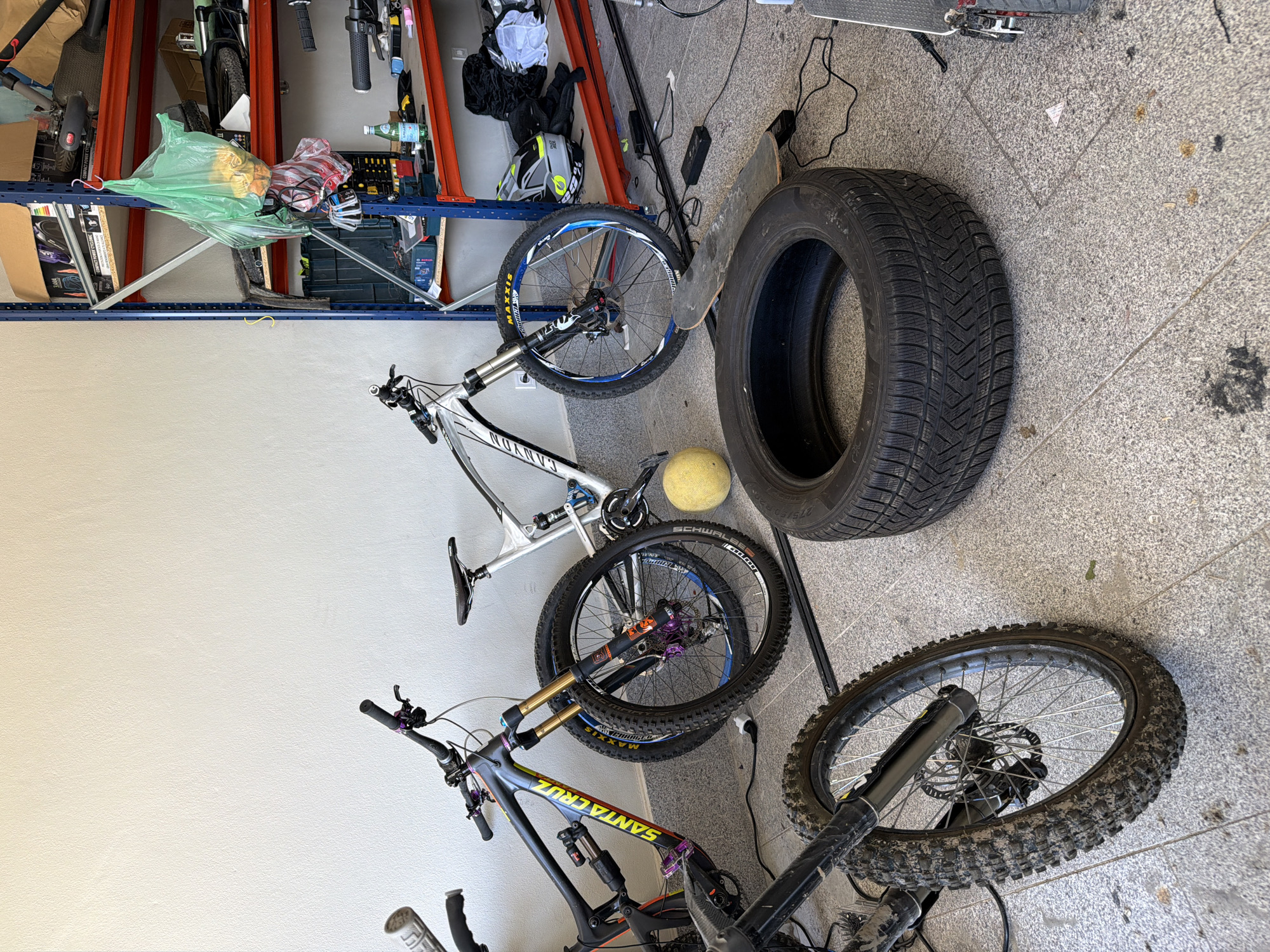 The image depicts a garage or storage area, likely in Asenovgrad, Bulgaria, containing bicycles, a car tire, shelving, and other miscellaneous items. Three bicycles are visible, including a Santa Cruz mountain bike, a Canyon mountain bike, and another mountain bike. The car tire is black and has a deep tread pattern. The shelving unit is made of metal and contains various items, including tools, bags, and containers. The floor is tiled with light-colored concrete squares. There are no people visible in the image. Other objects of note include a skateboard, a yellow ball, and various power cords. The environment suggests a residential or commercial space dedicated to storage and maintenance activities.
