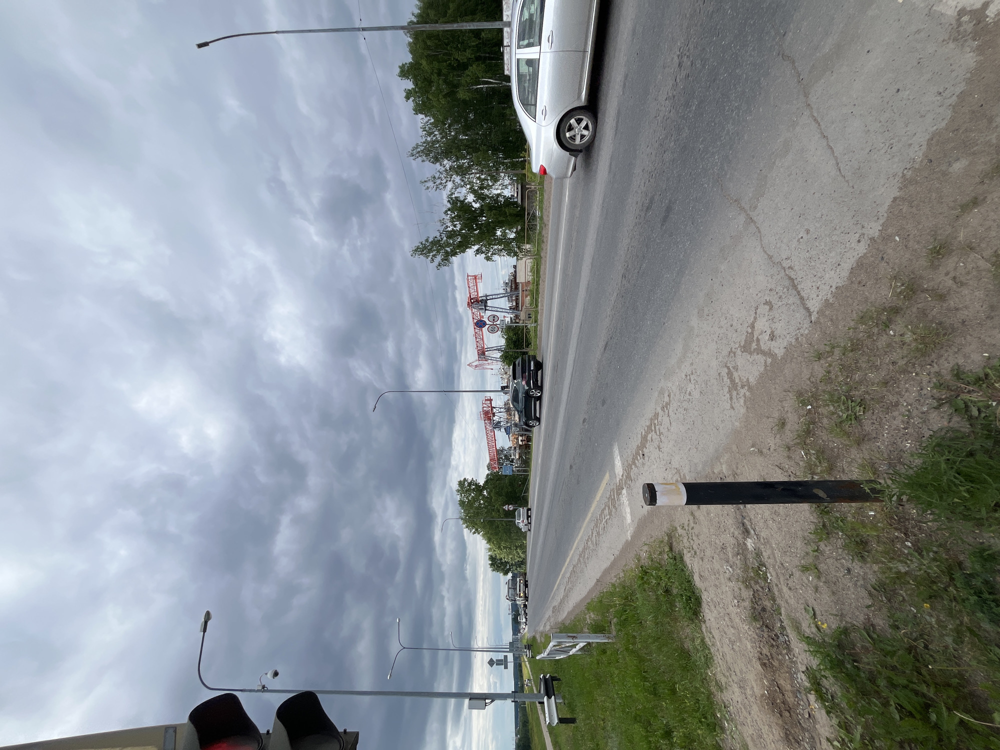 The image depicts a cloudy day on a two-lane asphalt road, likely in the city of Chaikovsky, Russia, given the context provided. The road is bordered by a grassy shoulder with a white metal fence on the left and a metal bollard. Several vehicles are visible on the road, including a silver sedan in the right lane and a black SUV behind it. In the distance, a bus and other cars are seen traveling in the opposite direction.
Overhead, a traffic light with a red signal is prominent on the left side of the frame, accompanied by a surveillance camera mounted on a pole. Further down the road, a series of large red industrial cranes are visible against the sky, suggesting proximity to a port or industrial area. Trees and vegetation line the right side of the road in the mid-ground. The sky is overcast with heavy, gray clouds.