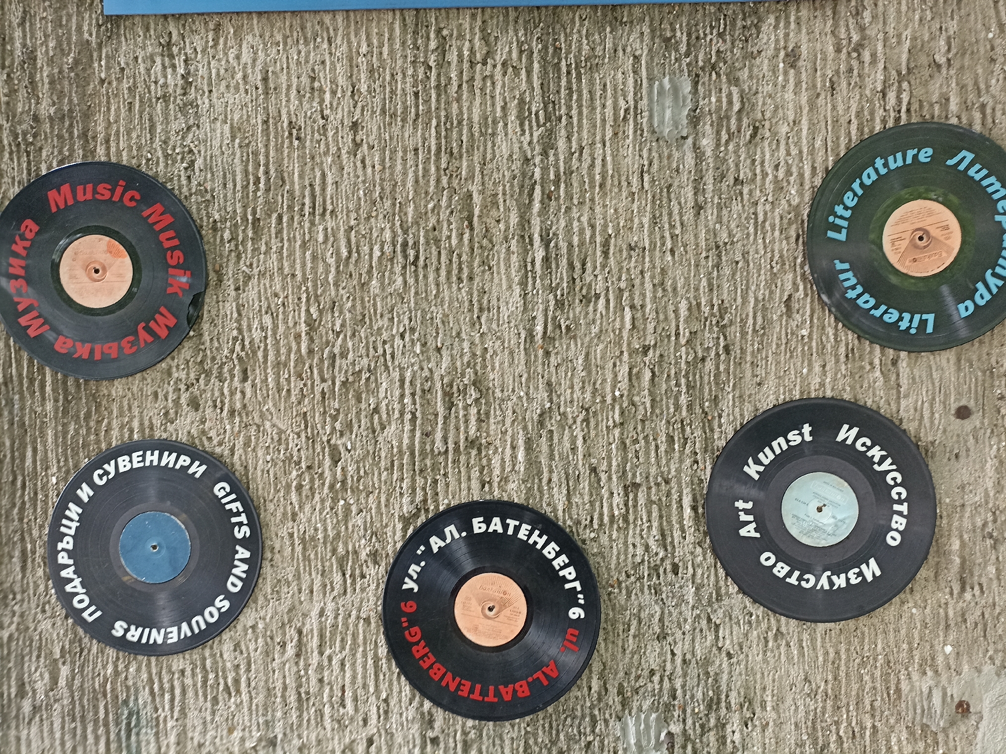 Five decorative discs resembling old vinyl records are affixed to a textured, light-colored concrete wall, likely a building facade in Burgas, Bulgaria. Each disc features text in multiple languages, including English, German, and Bulgarian (Cyrillic script), and revolves around a specific theme or information.
Starting from the top left and moving clockwise:
1. A black disc with red text features the words "Music," "Musik," and "Музика" (Music).
2. A dark green disc with light blue text displays "Literature," "Literatur," and "Литературa" (Literature).
3. A black disc with white text shows "Art," "Kunst," and "Изкуство" (Art).
4. A black disc with white and red text, indicating a street address: "ул. 'АЛ. БАТЕНБЕРГ' 6" and "AL. BATTENBERG" 6 (Al. Battenberg Street 6).
5. A black disc with white text reads "GIFTS AND SOUVENIRS" and "ПОДАРЪЦИ И СУВЕНИРИ" (Gifts and Souvenirs).
The discs have different colored central labels, some orange/brown, others blue. The wall itself is rough, with prominent vertical striations, and shows minor signs of wear or peeling in places. The overall display serves as an informational or decorative element on the street, likely pointing to nearby attractions, services, or themes relevant to the location. The presence of the street address "Al. Battenberg 6" directly links the display to a specific location within Burgas.
