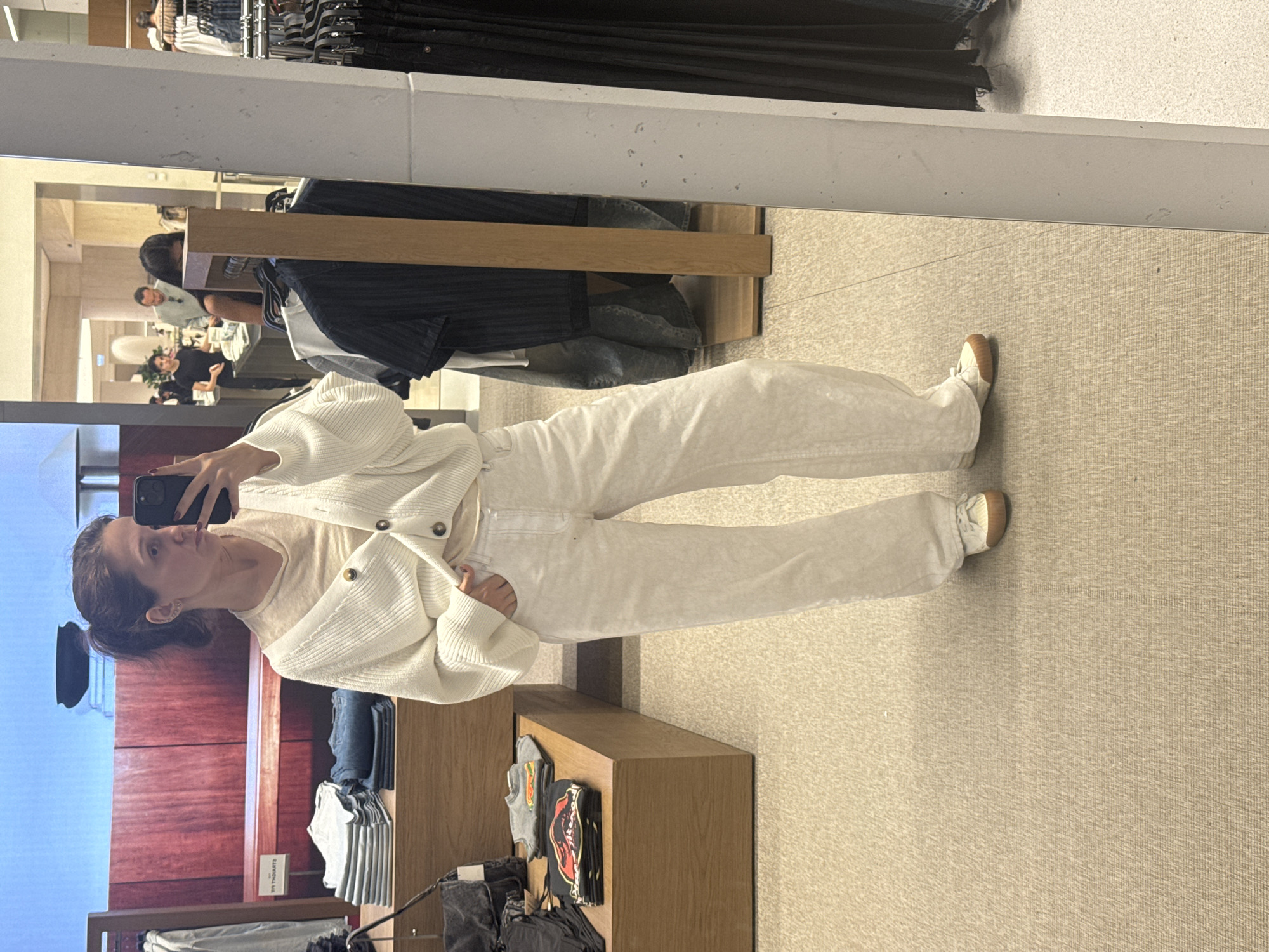 A woman is taking a mirror selfie in a retail store. She's wearing a white cardigan, a light-colored shirt, and white jeans with tan shoes. Behind her is a clothing rack with various items. In the background, people are visible in the store. The scene appears to be inside a clothing store in Dubai, United Arab Emirates.