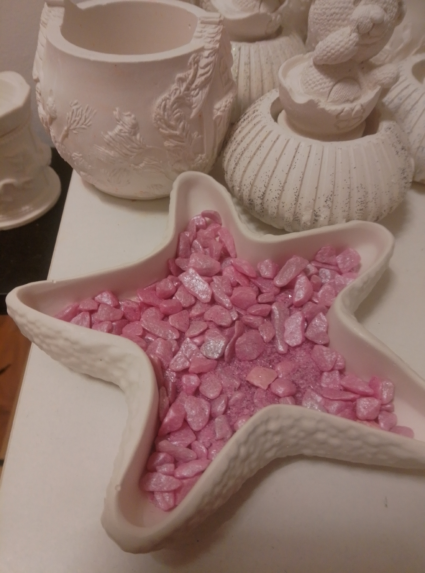 A close-up shot captures an arrangement of unpainted ceramic decorative items, possibly for crafting or display. In the foreground, a white ceramic dish shaped like a starfish is filled with small, pink, glittery stones. Behind the starfish dish, other ceramic pieces are visible. To the left, a cylindrical ceramic pot with raised decorative elements, resembling coral or foliage, stands. To the right and further back, a ceramic shell-shaped container with ribbed detailing and speckled with black glitter is partially visible. Another ceramic piece, possibly a small figure or object resting in a shell, is also present in the background. The lighting appears to be from overhead, casting soft shadows. The setting is indoors, likely on a white surface that could be a table or shelf. The image does not provide any information or context related to the city of Wedel, Germany.
