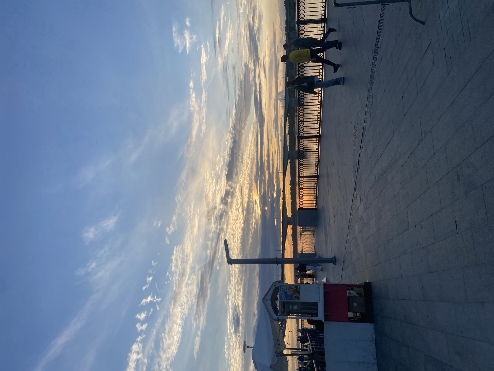 The image shows a sunset over the Kama River embankment in Perm, Russia. A wide paved walkway runs along the river, lined with a decorative black metal fence. Several people are strolling along the walkway, silhouetted against the glowing sky and water. In the foreground on the left, there is a white kiosk with a red trim and an umbrella providing shade. A lamppost stands prominently on the walkway. In the distance, a bridge spans the river, and the setting sun casts a warm golden light across the water and the sky, which is dotted with wispy clouds. The overall atmosphere is peaceful and serene, capturing a moment of evening relaxation by the river.