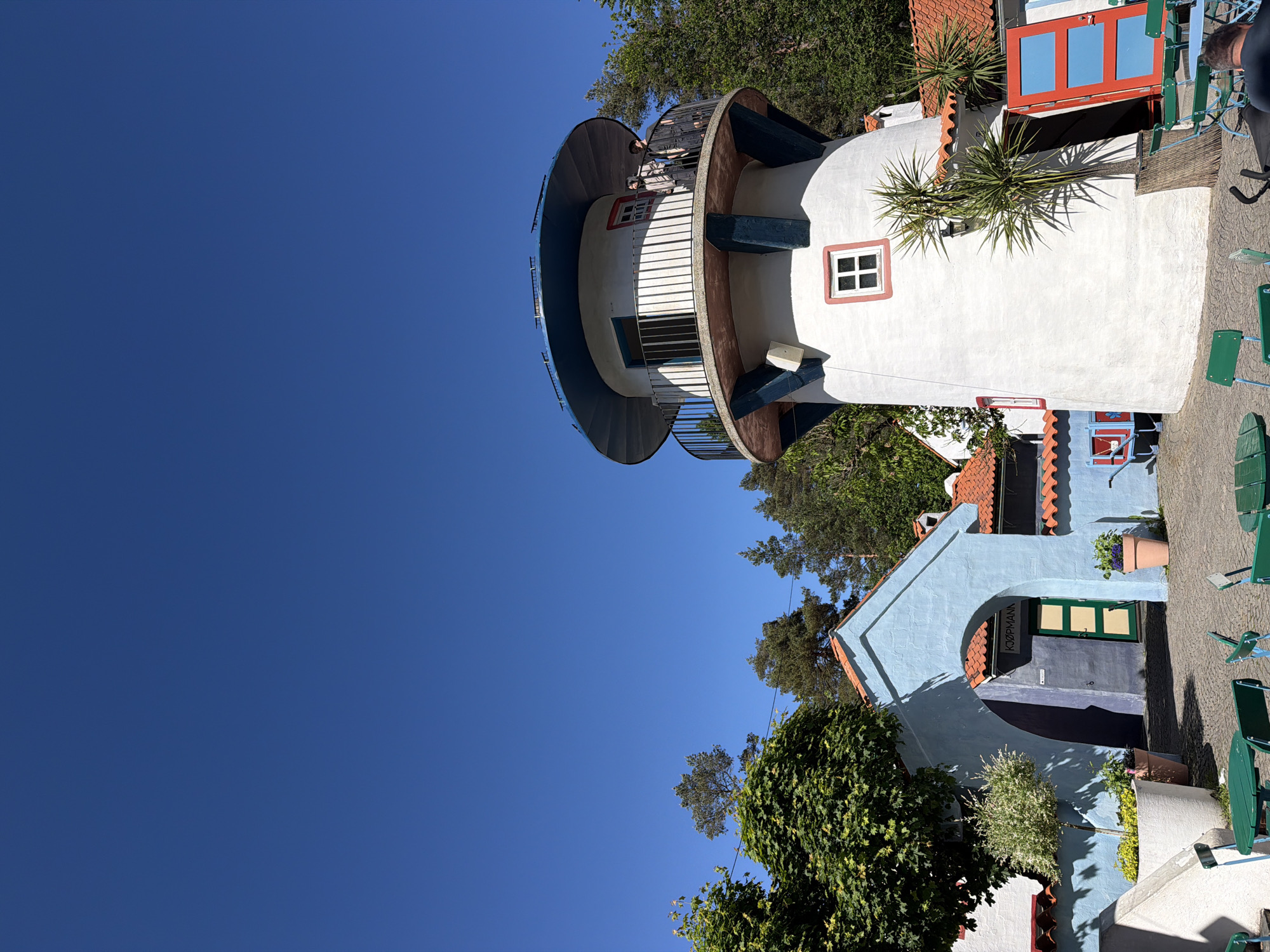 The image depicts an outdoor scene in Kristiansand, Norway, under a clear blue sky and bright sunlight. The central features are a white, cylindrical, lighthouse-like structure and a light blue archway building, both characterized by red-tiled roof sections.
The lighthouse-like structure, positioned on the right, features a white stucco-like exterior with a square window framed in white with red trim on its lower section. Above this, a prominent dark blue, circular platform is supported by blue-painted wooden beams. A balustraded balcony with a black metal railing wraps around the upper part of the structure, where a blue-framed door is visible. Two individuals are present on this balcony; one, dressed in a light-colored jacket, stands near the railing, partially obscured, while another, in a dark jacket, stands further back. A white rectangular utility box is affixed to the white wall below the balcony. To the right of the structure's base, a tall, potted plant with spiky leaves stands, adjacent to a vivid red double door with light blue panels.
On the left side of the image, a light blue building with a prominent archway leads into a darker blue interior where a green door is visible. A sign reading "KJØPMANN" is mounted above this door. Red-tiled roofs are evident on this building and others in the background. A large, mature tree with dense green foliage occupies the upper left, while a smaller, bushy plant in a white planter sits near the archway. Other potted plants are also visible.
The ground in the foreground is paved, and several green metal chairs and small round green tables are arranged, suggesting an outdoor seating area. The back of a person's head is visible in the bottom right, seated at one of these tables. The background features a dense collection of green trees, including both conifers and deciduous types, indicating a forested or natural environment surrounding the architectural elements. Strong shadows are cast due to the direct overhead sun.