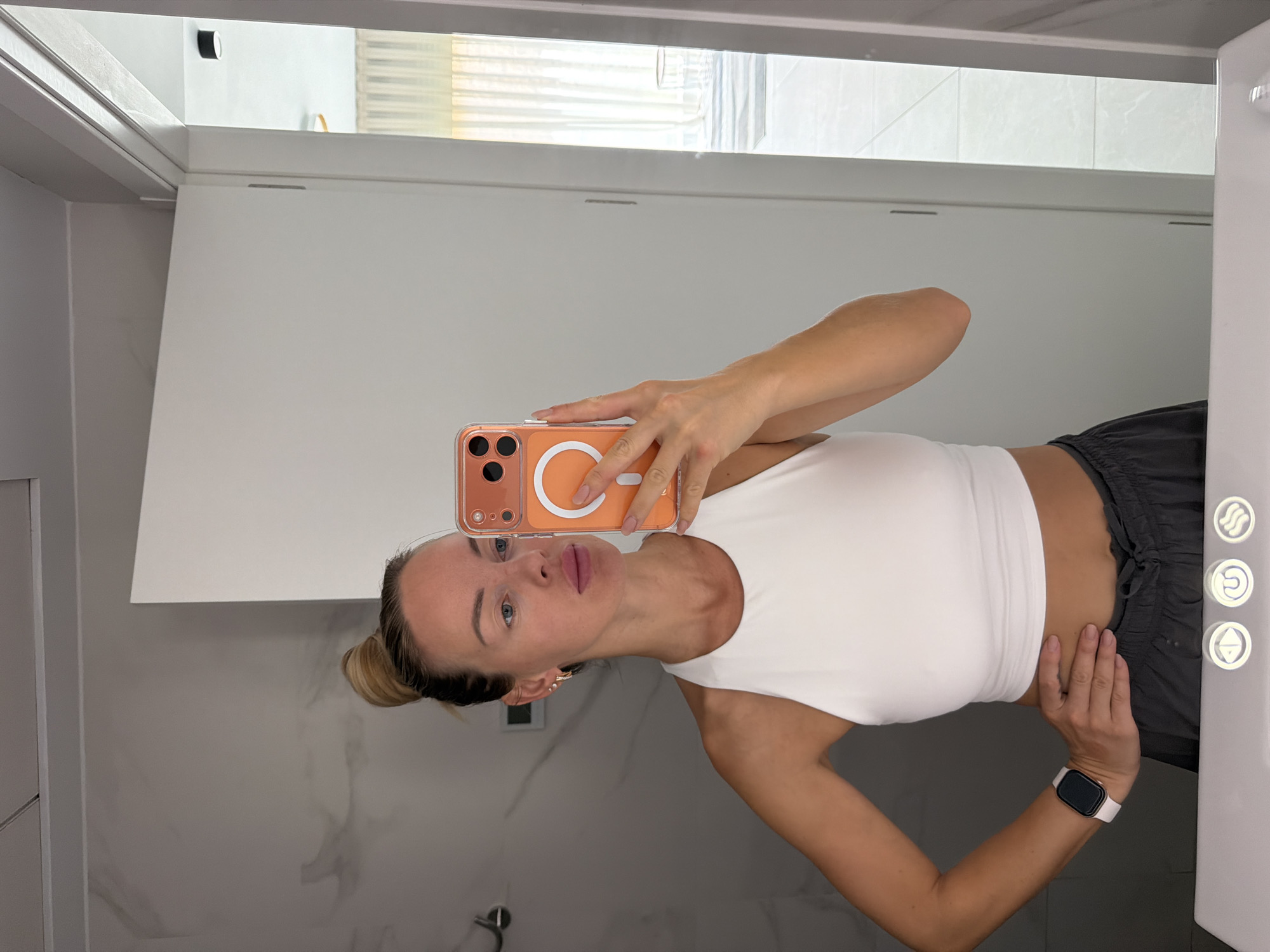 A woman with blonde hair in a bun takes a selfie in a modern bathroom. She is wearing a white crop top and dark grey shorts. A pink iPhone with an orange case is held up to take the photo. The bathroom has marble-patterned walls, a white door, and a white vanity with lights. In the reflection, a room with curtains and a chair can be seen. The location is identified as Finestrat, Spain.