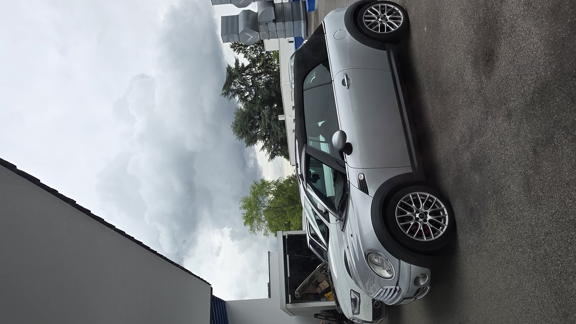 The image shows a silver convertible Mini Cooper parked outdoors on an asphalt surface, likely in a parking lot or a street. The car is the primary subject, with its top down, revealing a black interior. In the background, another white car is visible with its hood open, suggesting it may be undergoing maintenance. Further back, there are buildings and trees under a cloudy sky. To the right, a series of large, metallic structures resembling ventilation units or sculptures are visible. The overall scene appears to be an urban or industrial area, possibly a car service center or a commercial district.
