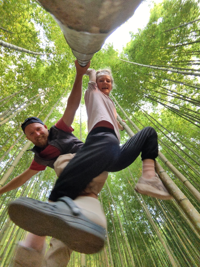 A low-angle photograph captured in a dense bamboo forest in Mù Cang Chải, Vietnam, shows two individuals, one male and one female, suspended by their hands from a thick, horizontal bamboo pole that crosses the upper portion of the frame. The male, positioned on the left, has a beard and wears a black cap, a dark vest over a red shirt, and tan pants; his left hand grips the pole, and he smiles towards the camera. The female, on the right, wears a light pink shirt, dark pants, and athletic shoes with pink socks; her right hand grips the pole, and she appears to be laughing or exclaiming, also looking towards the camera. Her legs are bent, with her body partially supported by the male. The background consists of numerous slender, green bamboo culms extending vertically towards a bright, hazy sky, characteristic of a bamboo grove.