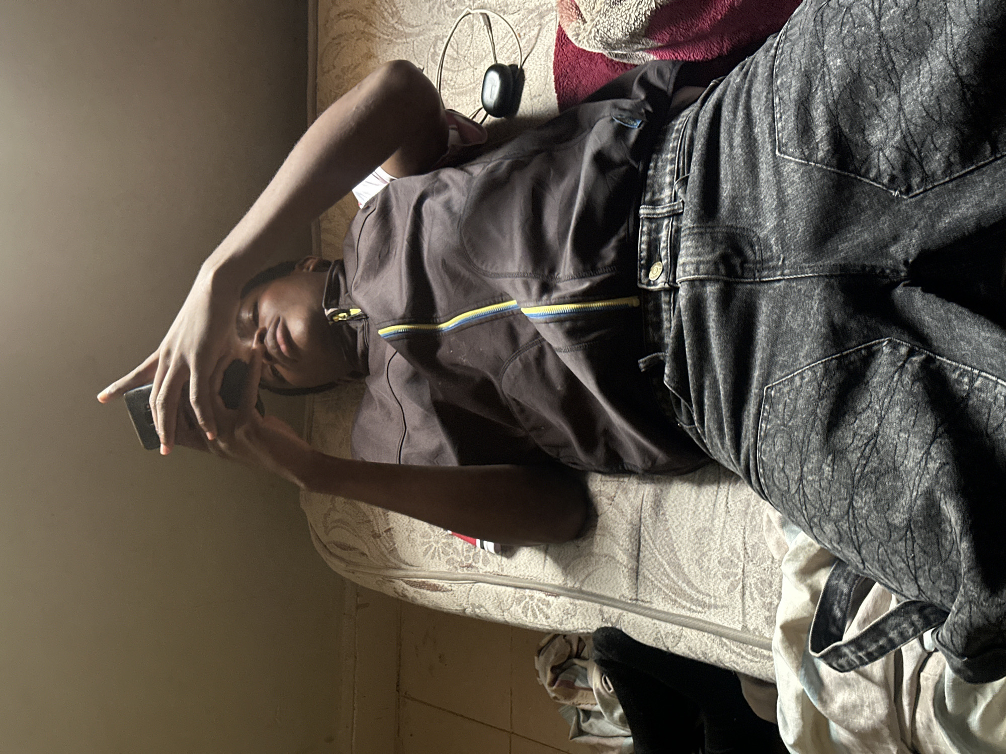 An individual, appearing young, lies supine on a light-colored, patterned mattress in a dimly lit indoor setting in Unknown, Unknown. The person, wearing a dark brown sleeveless jacket with a yellow and blue zipper and dark, possibly patterned or distressed, denim jeans, holds a black smartphone with both hands directly above their head, gazing at the screen. Orange nail polish is visible on the fingers. Adjacent to the person's right arm on the mattress, a black wireless earbud case, a white charging cable, and a segment of red fabric are present. The floor to the left of the mattress is light-colored and cluttered with various textiles. The surrounding walls and ceiling appear light brown or tan.