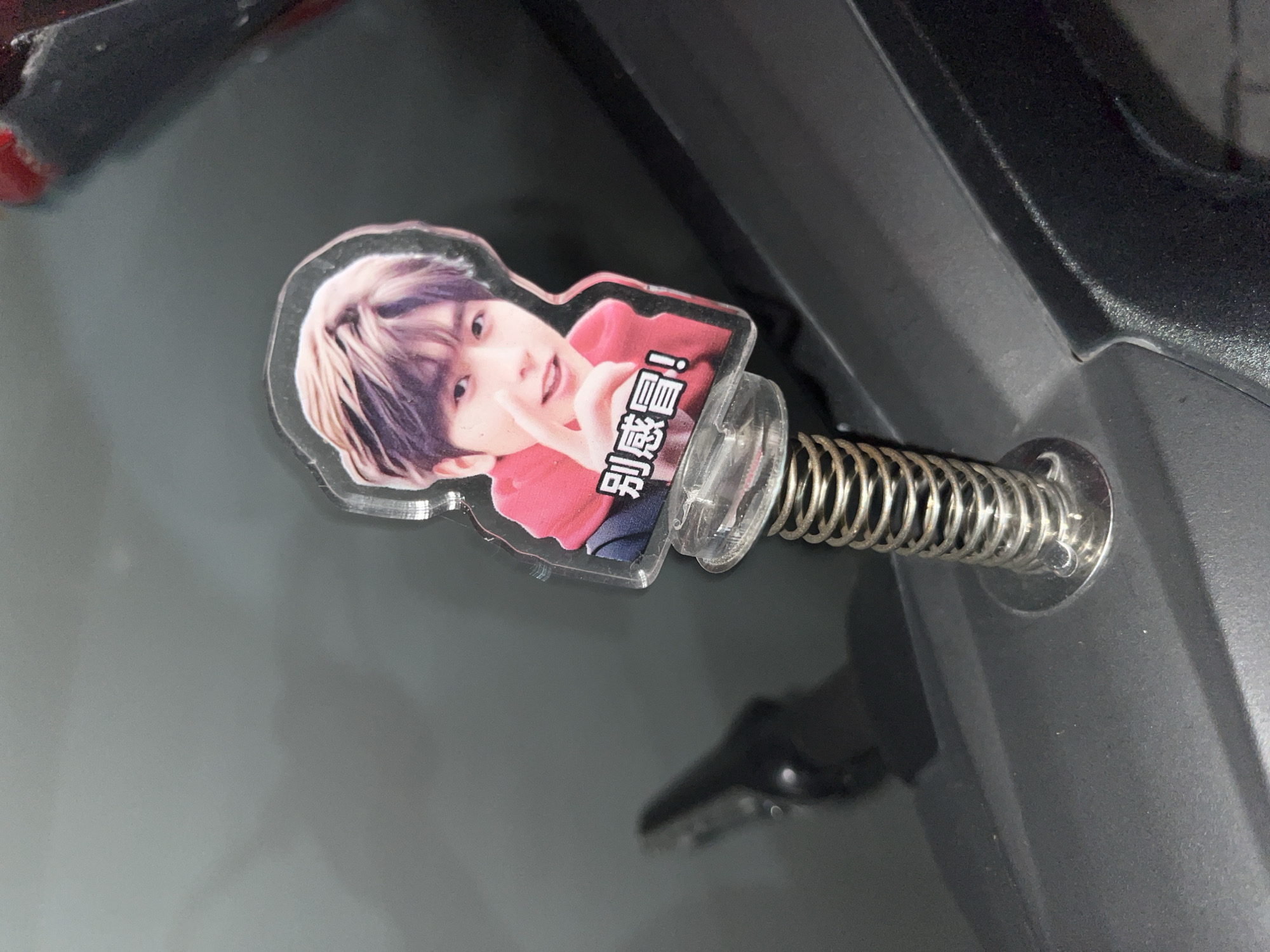 A small, plastic, cut-out figure of a young East Asian male with short, styled brown and blonde hair is attached to a metal spring. The figure is wearing a red hoodie. Text in Chinese characters, reading "别感冒!", is visible on the figure's clothing. The spring is attached to a metallic base that is affixed to a dark gray, textured surface, possibly the dashboard of a vehicle. The background is a blurred, dark gray, reflective surface. The scene appears to be indoors, and the object's placement suggests it is a car accessory.