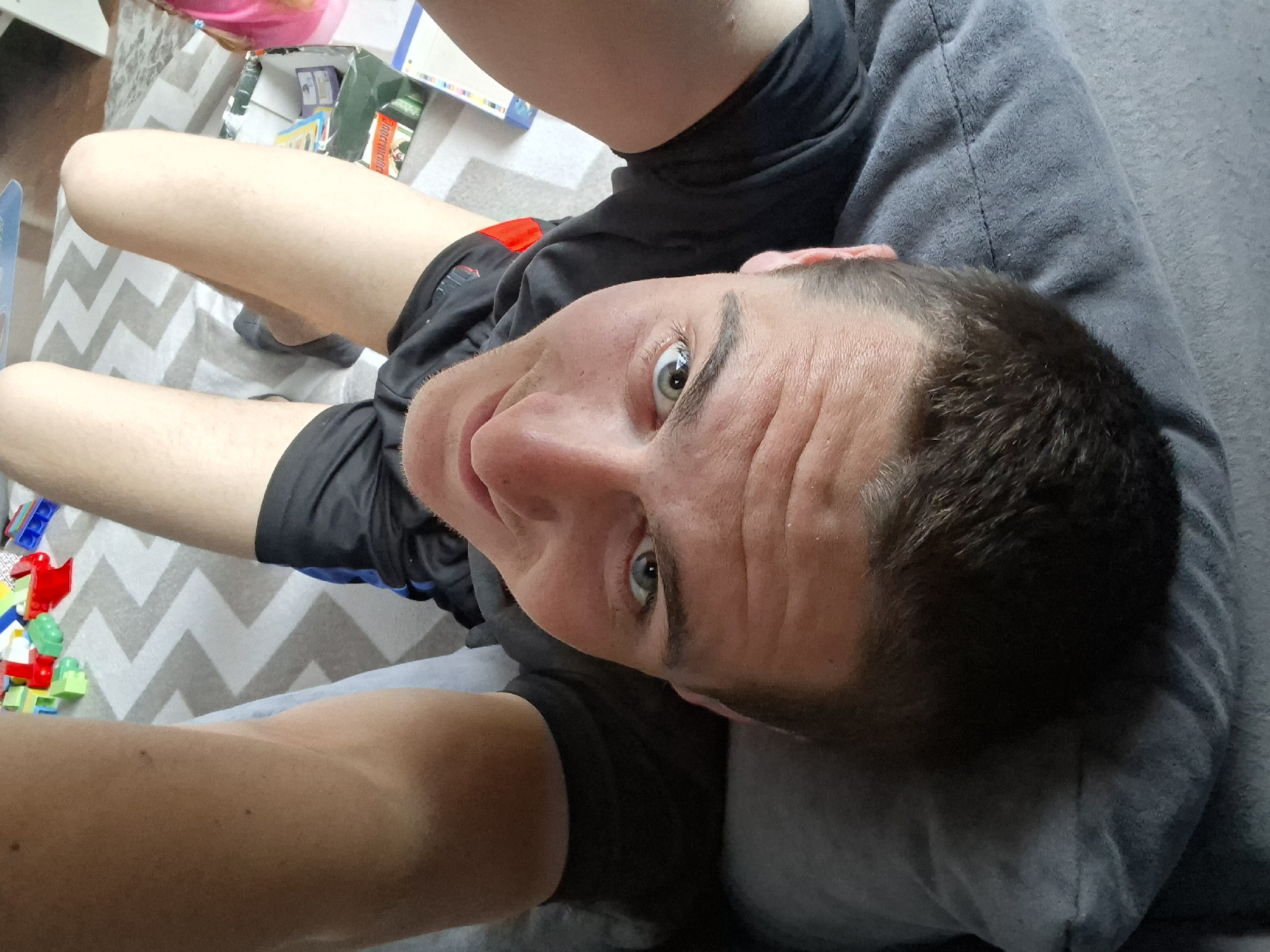 A man with short brown hair and gray eyes is lying on a gray pillow or cushion. He is wearing a black t-shirt and dark shorts with a red patch. His legs are visible, extending towards the top left of the frame. The man is looking up at the camera. On the floor, there is a gray and white zigzag patterned rug, along with various items including colorful plastic building blocks and boxes. The location is unknown.