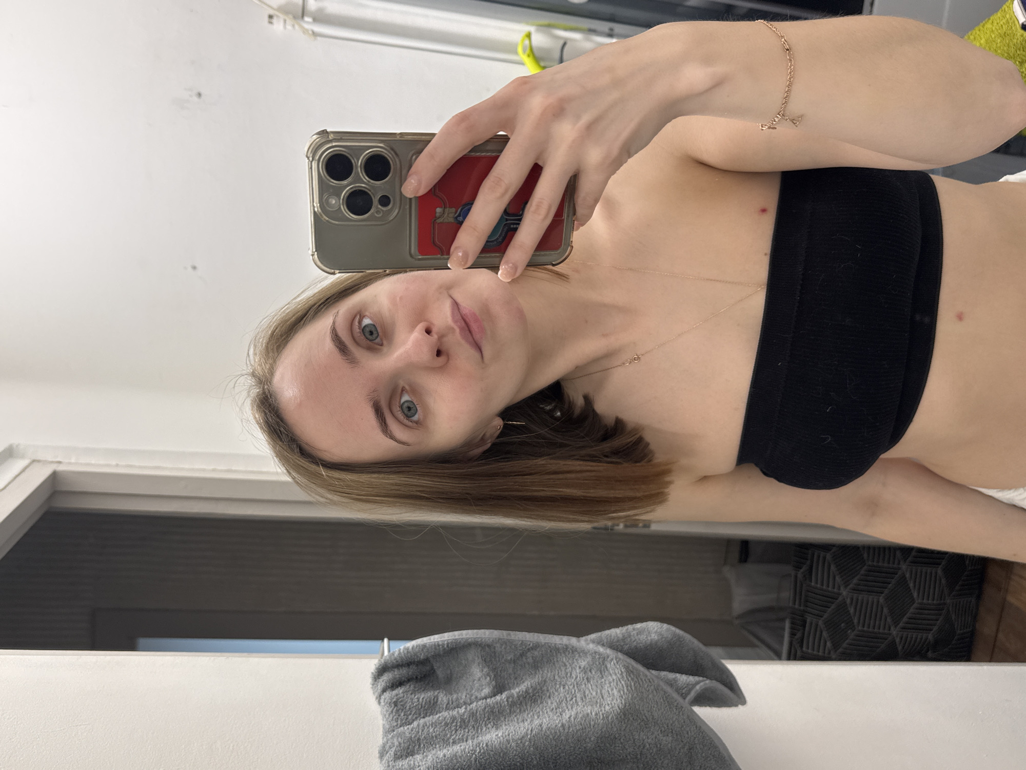 A young woman is taking a selfie in a bathroom. She is holding a phone with a case that appears to have a credit card visible through the back. The woman is wearing a black strapless top and a delicate gold necklace. A gray towel hangs near the lower part of the frame. The location is unknown and there's nothing to suggest the image was taken in Perm, Russia.