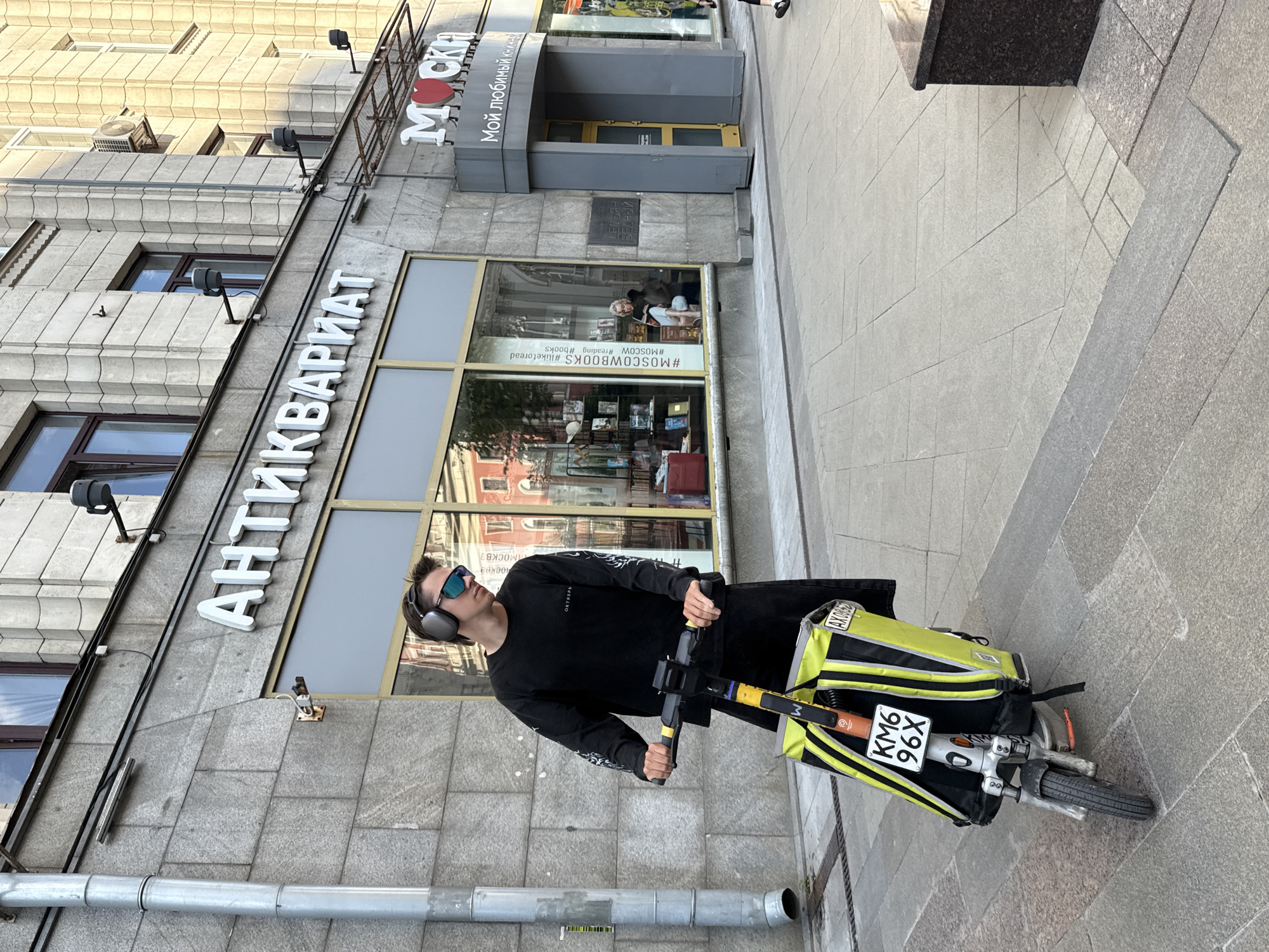 A young man wearing large over-ear headphones and sunglasses is standing on a sidewalk in Moscow, Russia, holding the handlebars of a silver electric scooter. The scooter has a bright yellow and black delivery bag attached to the front. The man is wearing a black long-sleeved shirt and black pants. Behind him is a building with large windows. The building has a sign that reads "АНТИКВАРИАТ" in large white letters. To the right of the "АНТИКВАРИАТ" sign is another sign with two red heart symbols and the word "МОСКВА" in large white letters. The sidewalk is made of grey paving stones, and there is a granite planter to the right of the image.