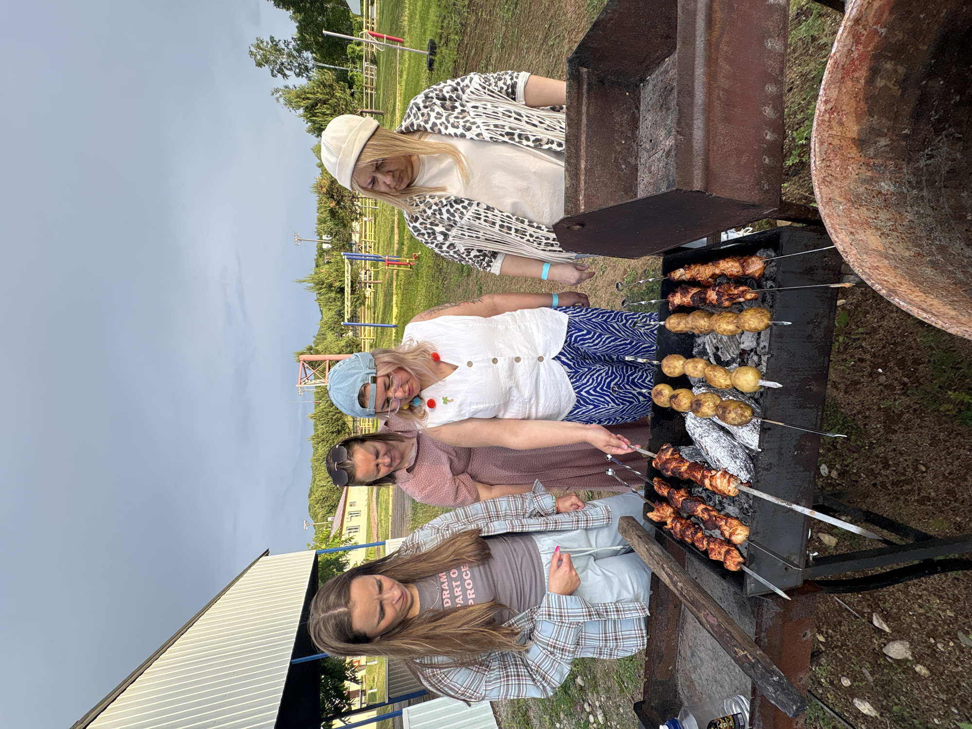 Four women are standing around a barbecue grill in an outdoor setting. The grill contains skewers of meat and potatoes, some of which are wrapped in aluminum foil. The woman in the foreground on the left is wearing a plaid shirt and light-colored pants. The woman next to her is wearing a pink dress and sunglasses. The woman in the center is wearing a white vest over a blue and white striped skirt and a blue baseball cap. The woman on the right is wearing a patterned jacket and a cream-colored hat. The background shows a building with a metal roof, trees, and a playground structure. The sky is overcast. The location is stated to be in the city of Birobidzhan, Russia.