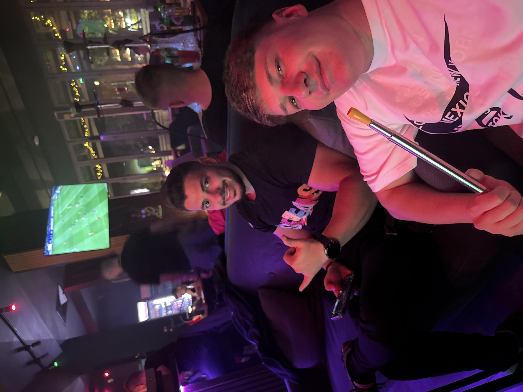 Two young men are seated closely together in a dimly lit establishment, possibly a bar or lounge. The man on the right is fair-skinned and wearing a white T-shirt with a black Nike swoosh and lettering. He holds a silver hookah mouthpiece. The man on the left has dark hair and a beard, and he is wearing a dark T-shirt. Both men are smiling at the camera. In the background, there are other people, a television screen displaying a soccer game, and decorative lights. Through a window, there's a glimpse of foliage and city lights. The location is likely in Plovdiv, Bulgaria, given the request. The scene appears relaxed and social.