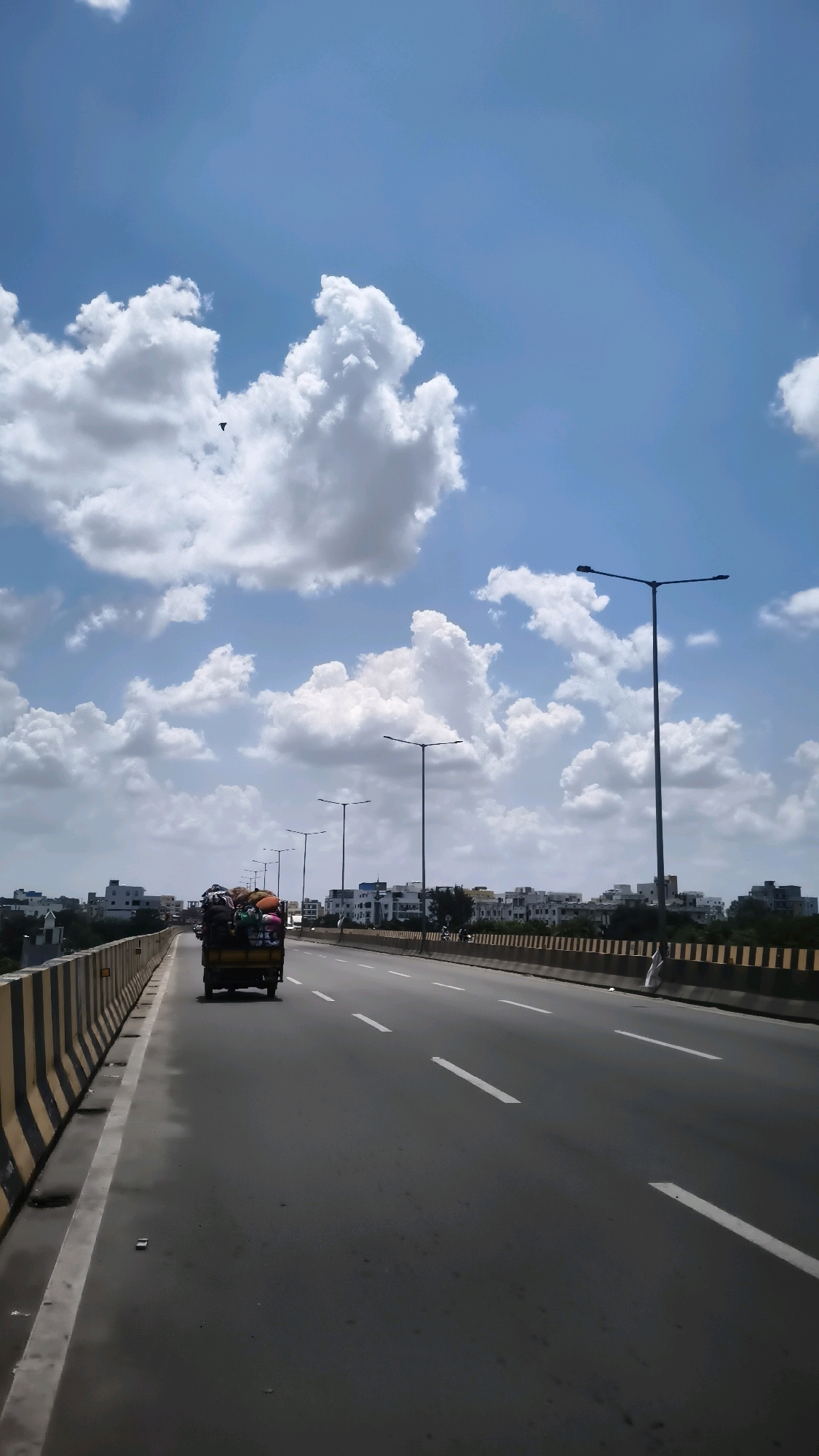 The image shows a clear sky with fluffy white clouds over a highway in Hyderabad, India. A truck, laden with various items covered with tarpaulin, is traveling in the left lane. The highway is lined with streetlights and a yellow and black striped barrier on the left. Buildings are visible in the background on the right side of the highway. The road has white lane markings. A bird is visible in the sky.