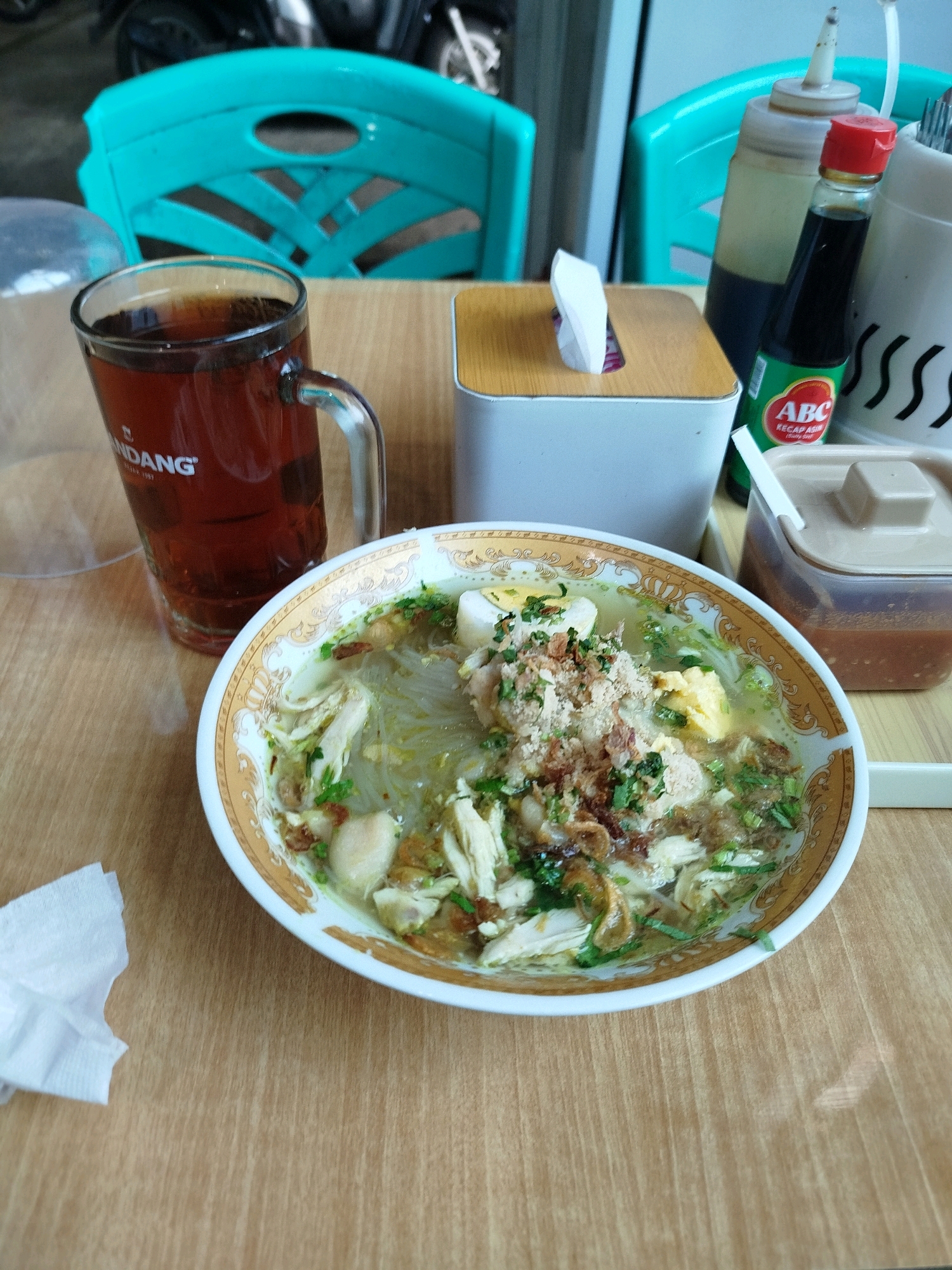 The image shows a close-up, eye-level view of a meal setting on a wooden table. In the foreground, a white napkin is crumpled on the left. To the right, a bowl of soto ayam, a chicken noodle soup, is prominent. The soto contains chicken pieces, noodles, boiled egg slices, and garnished with chopped green herbs and fried shallots. The bowl has a decorative gold rim. Behind the soup, a glass mug filled with dark tea is visible, with the word "UNDANG" partially visible on its side. To the right of the soup and tea, a tissue box with a light brown wooden lid sits on a white rectangular container. Next to it, a bottle of ABC Kecap Asin (sweet soy sauce) with a red cap and green label is standing upright, next to a white squeezy bottle. Behind these items, a translucent plastic container with a brown lid holds a reddish sauce. In the background, two turquoise plastic chairs are visible, suggesting an outdoor or semi-outdoor dining area. The overall setting appears to be a casual eatery, possibly a warung or a small restaurant. The lighting is natural, coming from above and slightly to the right, casting a subtle shadow on the table. The environment suggests a warm climate, and the items on the table are typical of Indonesian cuisine. The location is likely a food stall or restaurant in Balikpapan, Indonesia, given the presence of typical Indonesian food and condiments.