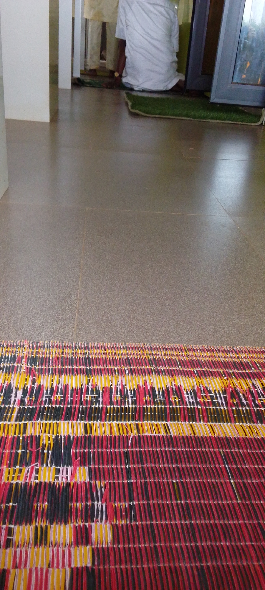 The image shows a tiled floor with a colorful mat in the foreground. In the background, two people are sitting on a green mat near a window. The scene appears to be indoors, possibly in a religious building or community center. It is impossible to confirm that this image was taken in Jos, Nigeria, based on visual information alone.