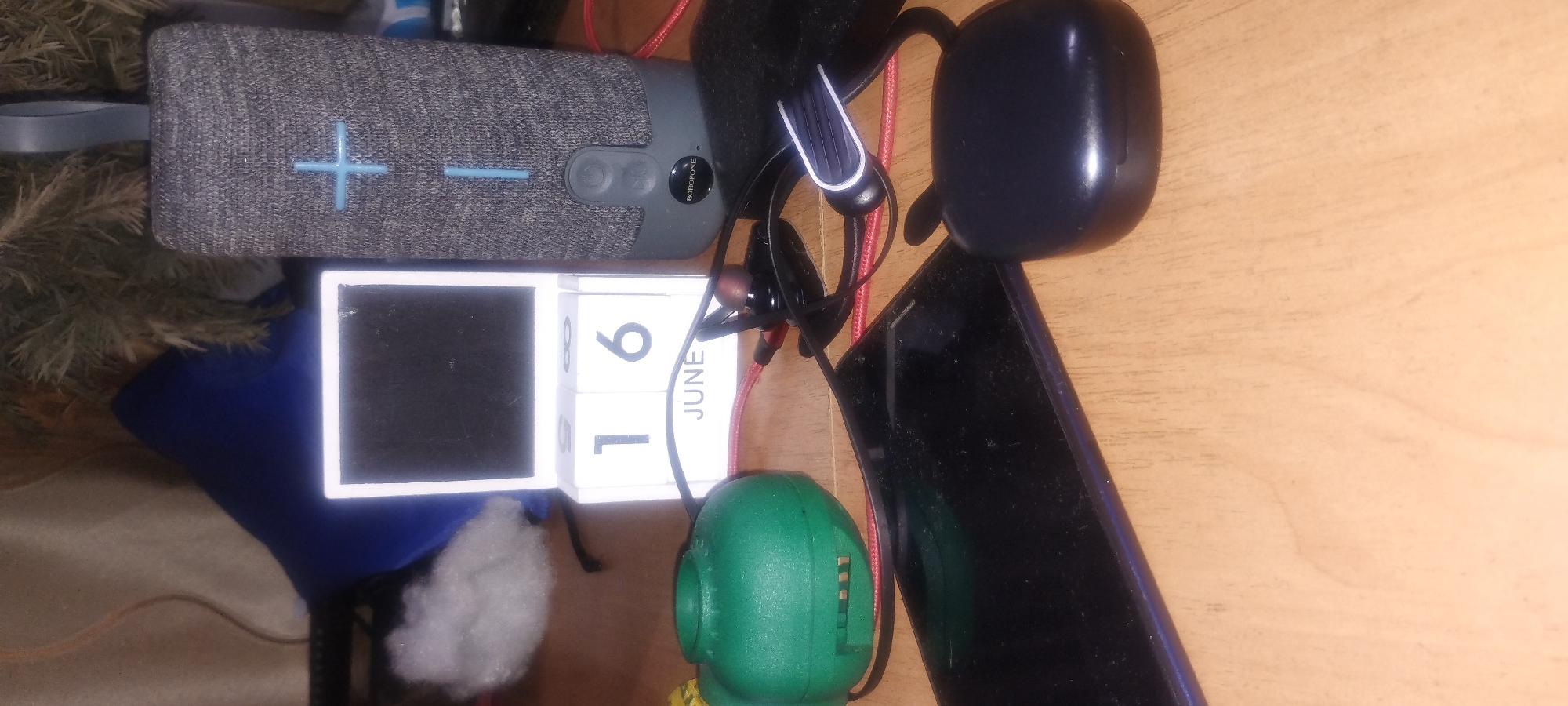 The image shows a collection of electronic devices and a decorative calendar arranged on a wooden surface, likely a table. In the background, out of focus, is a blue pillow and some greenery, possibly a plant or tree branches.
The prominent objects are:
* A cylindrical Bluetooth speaker, gray with blue accents, displaying a plus-sign symbol and a button labeled "BOROFONE."
* A small, white calendar block showing "16" on one cube and "JUNE" below.
* A dark-colored smartphone lying flat on the table.
* A pair of black earbuds with red detailing, connected by wires.
* A bright green, dome-shaped object, possibly a small container or toy, with slits on its side.
* A black charging case for earbuds situated to the right of the smartphone.
The lighting appears to be artificial and somewhat dim, casting shadows on the wooden surface. The overall scene suggests a personal workspace or living area. There are no people or animals present in the image. The location is unknown.