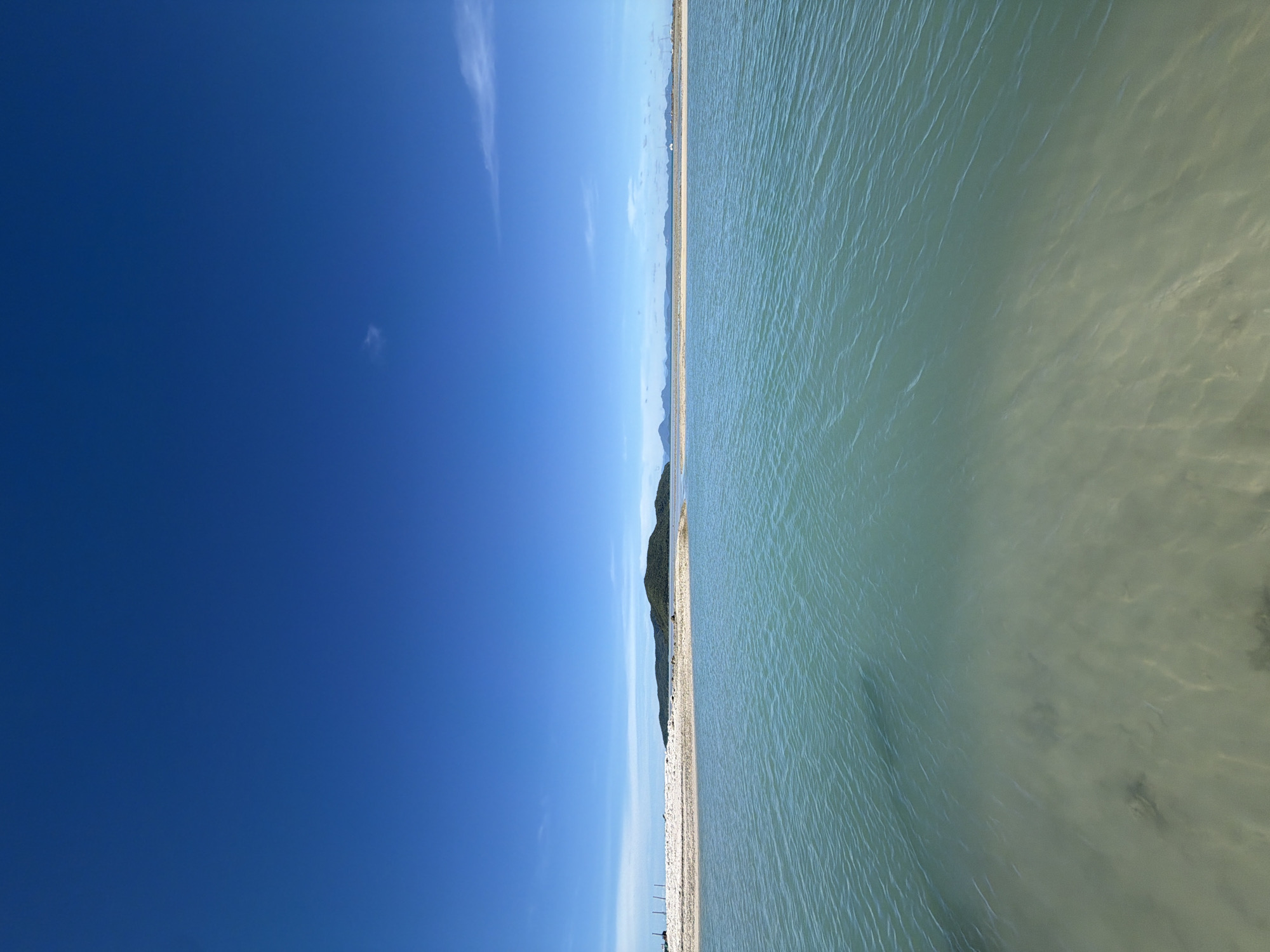 The image depicts a serene coastal scene in Baan Thong Krut, Thailand. The foreground is dominated by clear, turquoise water, revealing a sandy bottom with subtle ripples and occasional shadows. A narrow strip of white sand beach stretches horizontally across the midground, punctuated by sparse vegetation and a few vertical structures on the far left, possibly pilings or small poles. Beyond the beach, a verdant, tree-covered island rises, its silhouette sharp against the horizon. In the distance, a chain of hazy mountains forms a backdrop to the tranquil seascape. The sky above is a vibrant, cloudless blue, with a single wisp of white cloud visible near the top center. The overall impression is one of peaceful isolation and natural beauty.