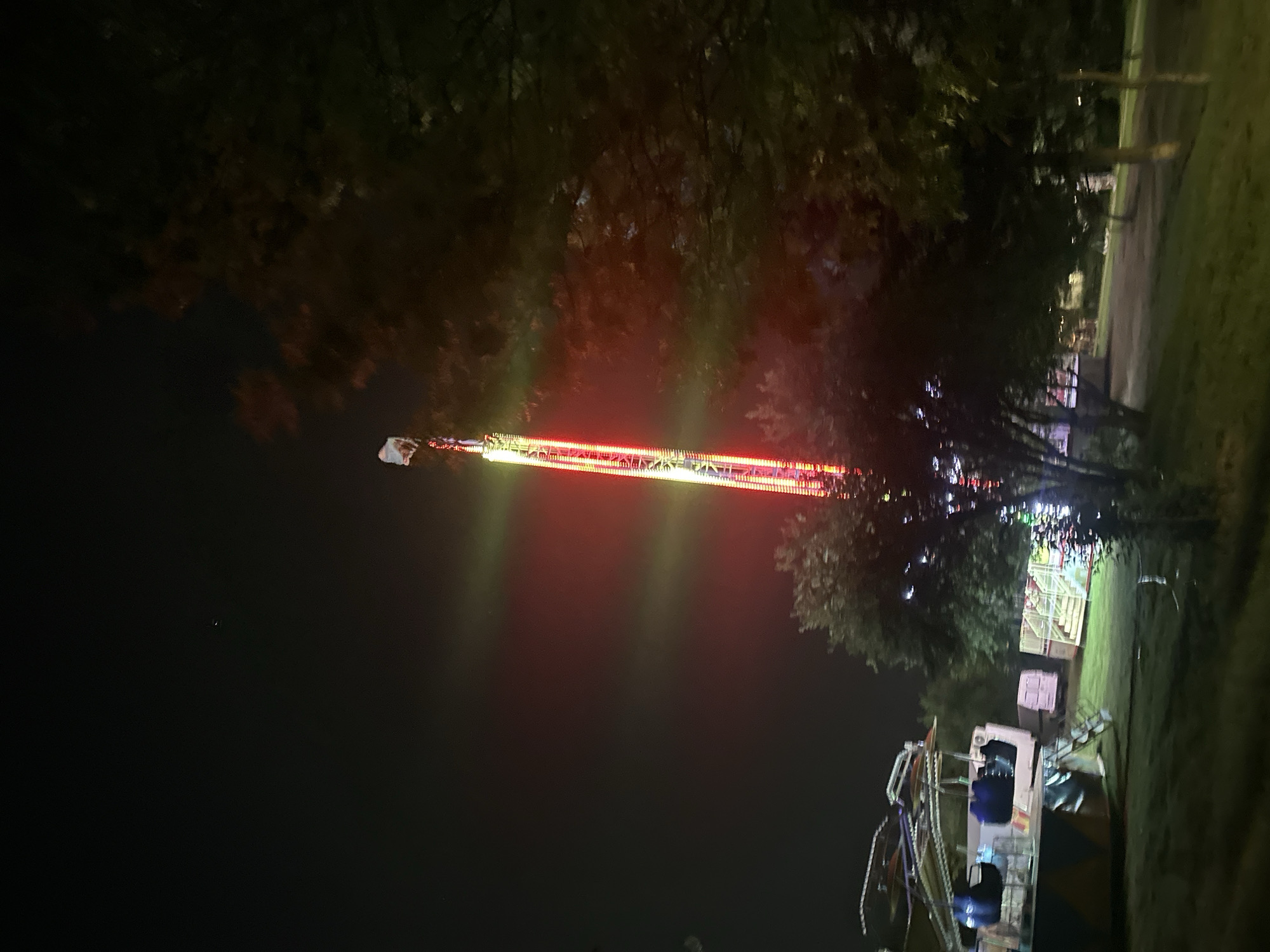An outdoor fairground scene at night features a tall, illuminated amusement park ride that is red and white. To the left of the ride, several other carnival attractions are visible, including a ride with suspended seats and a pink trailer. Trees are scattered throughout the scene, some of which are illuminated by the lights of the fairground. The ground is covered in grass. The sky is dark and shows a few stars.
