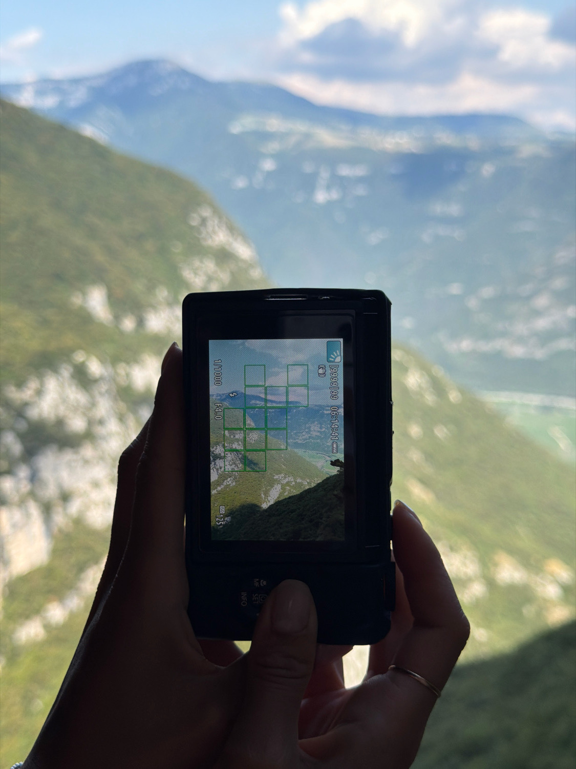 A hand, adorned with a thin gold ring on the ring finger, holds a black digital camera in front of a blurred, expansive mountain landscape. The camera's screen displays a grid overlaying a view of the same mountain vista, indicating it is actively framing a shot. The background features rolling green hills and distant, hazy mountains under a bright sky with scattered clouds. The scene suggests a moment of capturing the natural beauty of the mountainous terrain, likely in a location like Ferrara di Monte Baldo, Italy, known for its scenic views.