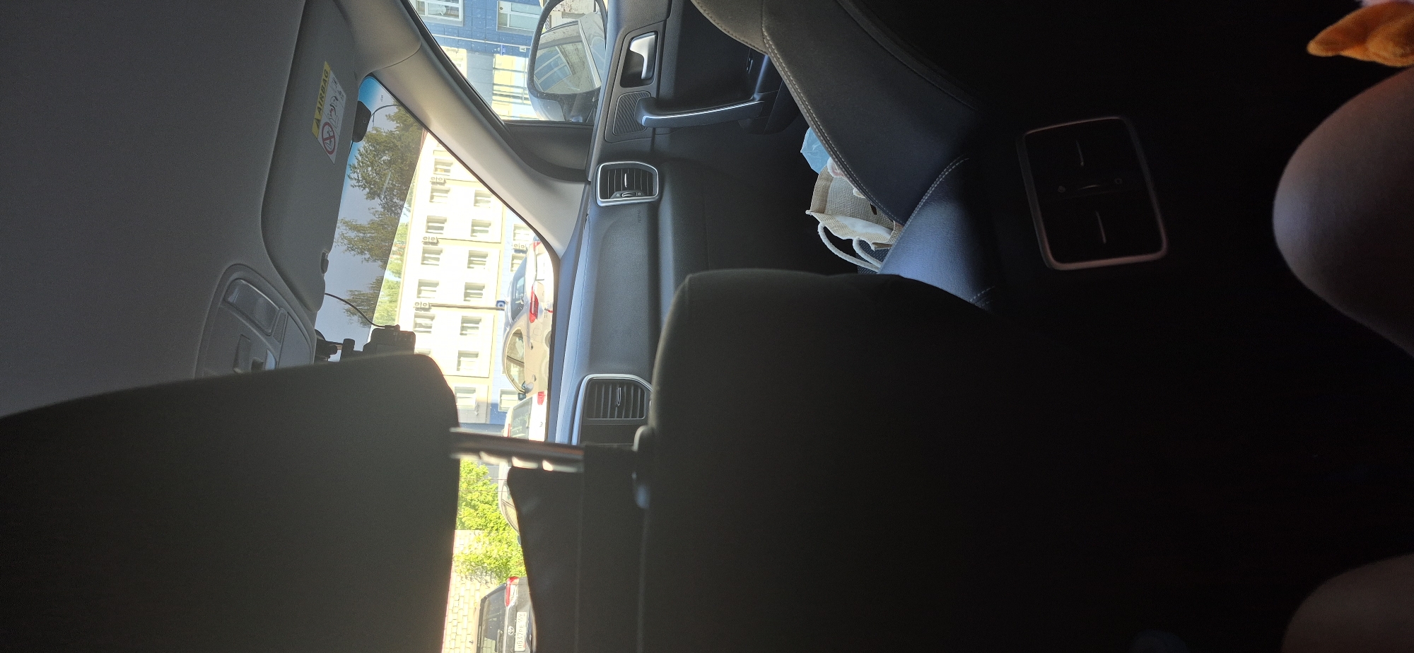 The image depicts the interior of a car, as viewed from the back seat. A partially visible driver's side window shows a cityscape with a light-colored building with multiple windows. The front passenger seat is black leather with adjustable vents, and behind it is a black leather seat. A mask is lying on the passenger seat. The details suggest the photo was taken during daytime, but the exact location is not discernable from the given information.