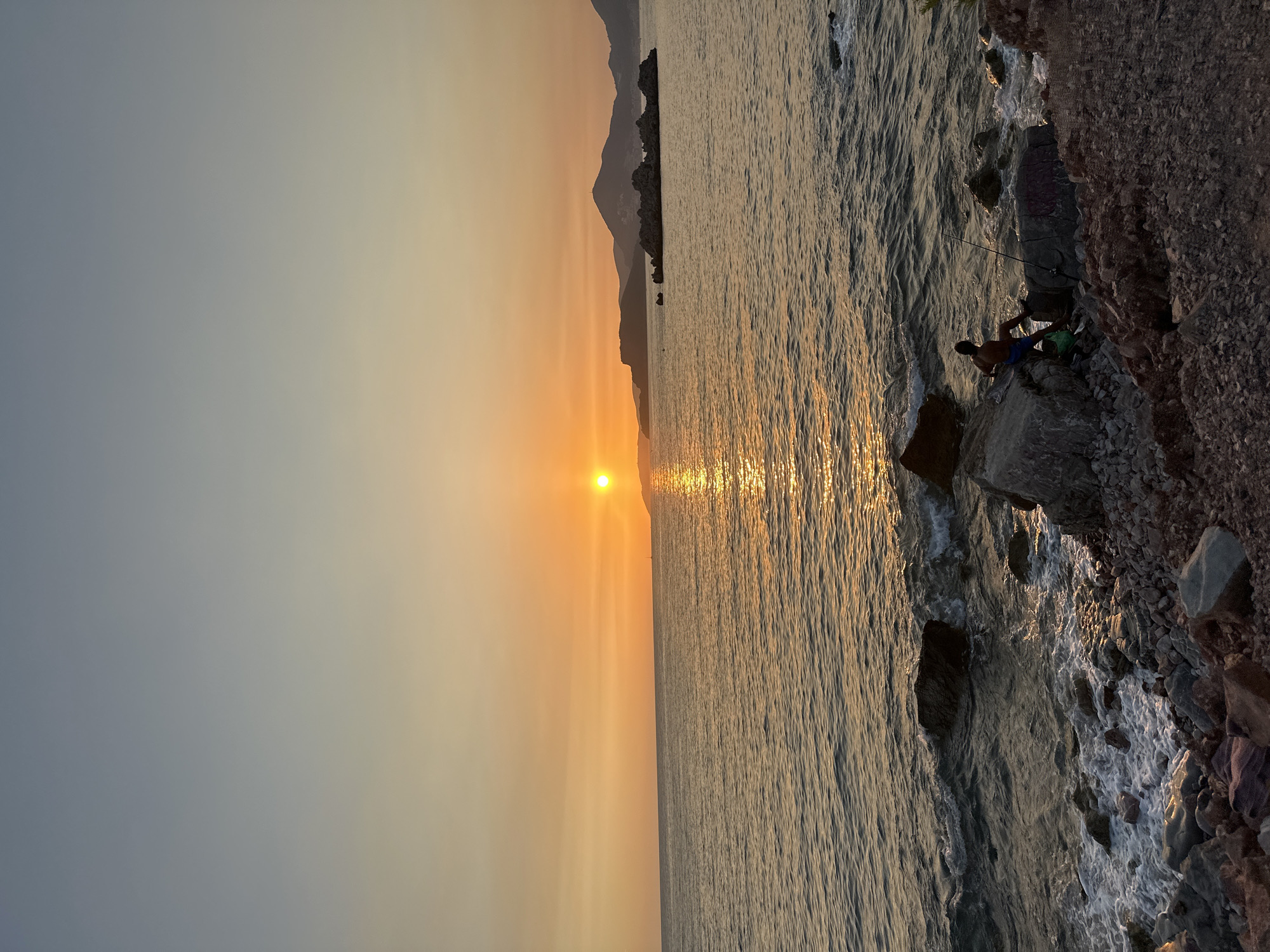 This image depicts a sunset over the Adriatic Sea, with Sveti Stefan, Montenegro, visible in the distance. The sun is a bright orange orb positioned low in the sky, casting a shimmering golden reflection on the water. The sea is moderately calm, with gentle waves lapping against a rocky shoreline in the foreground.
A lone figure, a man, is seen sitting on a large rock near the water's edge, engaged in fishing with a rod. He is wearing blue shorts and is partially obscured by the rock. Beside him, on the rock, is a green bag. The shoreline is composed of a mix of large and small rocks, with some vegetation visible on the far right.
In the middle ground, a cluster of rocky islets breaks the surface of the water, and further out, the mountainous coastline of Sveti Stefan is silhouetted against the orange sky. The sky transitions from a pale blue at the top to a warm orange hue near the horizon, indicating the late stages of sunset. The overall atmosphere is serene and tranquil.