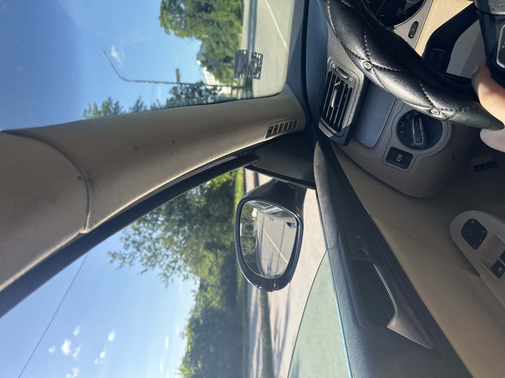 The scene depicts the interior of a car, specifically from the driver's perspective, looking forward and to the left, in the city of Gabrovo, Bulgaria. The time of day is sunny, with a clear blue sky and scattered white cumulus clouds.
Inside the car, the right hand of the driver grips a black, quilted, studded steering wheel cover. The dashboard is visible, featuring air vents and a rotary headlight switch set to the 'Auto' position on the left. The A-pillar and door panel on the driver's side are primarily a light beige or tan color, contrasted by darker grey/black elements such as the upper door trim, door handle, and window control panel. The side mirror on the driver's door reflects a segment of a paved road extending into the distance, bordered by green foliage.
Outside the car, through the windshield, a two-lane asphalt road is visible. The roadside is densely lined with green trees and bushes, extending into the background. A tall, curved street light pole stands on the right side of the view. In the far distance, behind the trees, a light-colored building is partially visible. Reflections on the windshield on the right side show what appears to be a dark rectangular sticker or vignette. The environment suggests an urban or suburban setting with ample greenery.
