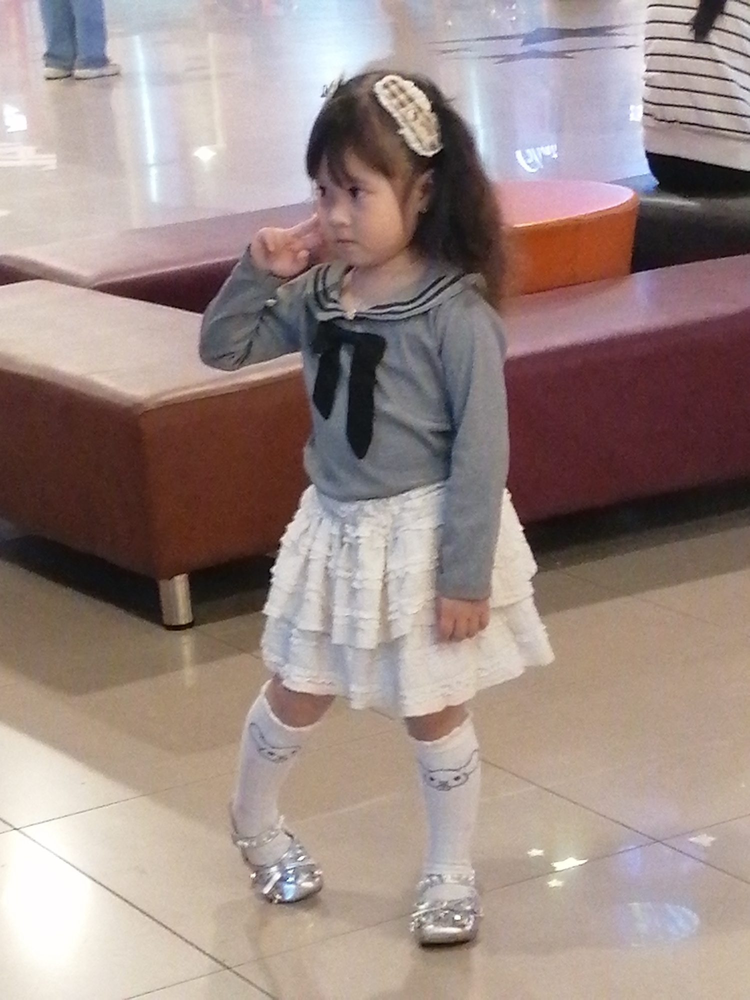 A young girl with dark hair, wearing a grey long-sleeved shirt with a black bow and a white tiered skirt, stands in what appears to be an indoor setting, possibly a mall or a large public space. She has a hair clip on the right side of her head and is wearing white socks with a cat design and silver shoes. Her left hand is raised to her face, with her index finger pointing towards her temple, as if in thought or making a gesture. The floor is tiled with a light-colored pattern. In the background, there are reddish-brown benches or seating areas, and beyond that, a person in blue jeans is visible, partially out of focus. A small orange circular object is also present in the background. The lighting suggests it is daytime, but the exact time is not discernible. The overall impression is of a candid moment captured while the girl is posing or playing. There is no visible text in the image.