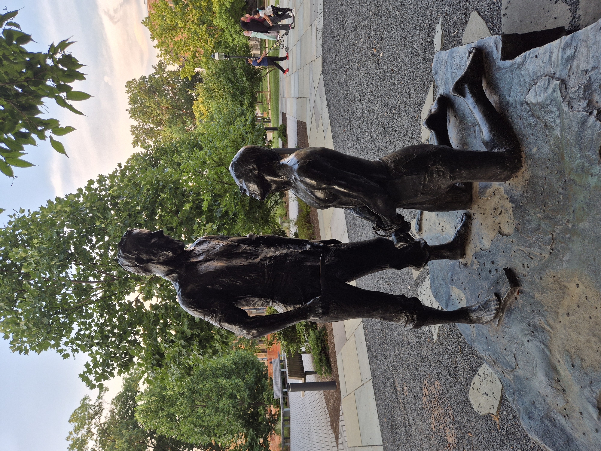 The image depicts a bronze sculpture of two men in an outdoor setting. The sculpture is positioned on a rough, dark stone base. One man stands with his hands in his pockets, looking down at the other man who is kneeling. The kneeling man has his head tilted back and his hands appear to be bound together in front of him. Both figures are muscular and appear to be wearing rough clothing, possibly representing laborers or historical figures.
The sculpture is located in Princeton, New Jersey, USA, in a plaza area. Behind the sculpture, there is a paved walkway with a patterned brick design. Several benches are visible in the background, along with lush green trees. In the far background, to the right of the sculpture, several people are walking and one individual is on a scooter. The sky is visible above, with a soft blend of orange, pink, and blue hues, indicating it might be either dawn or dusk. The ground surrounding the sculpture is covered in dark gravel.