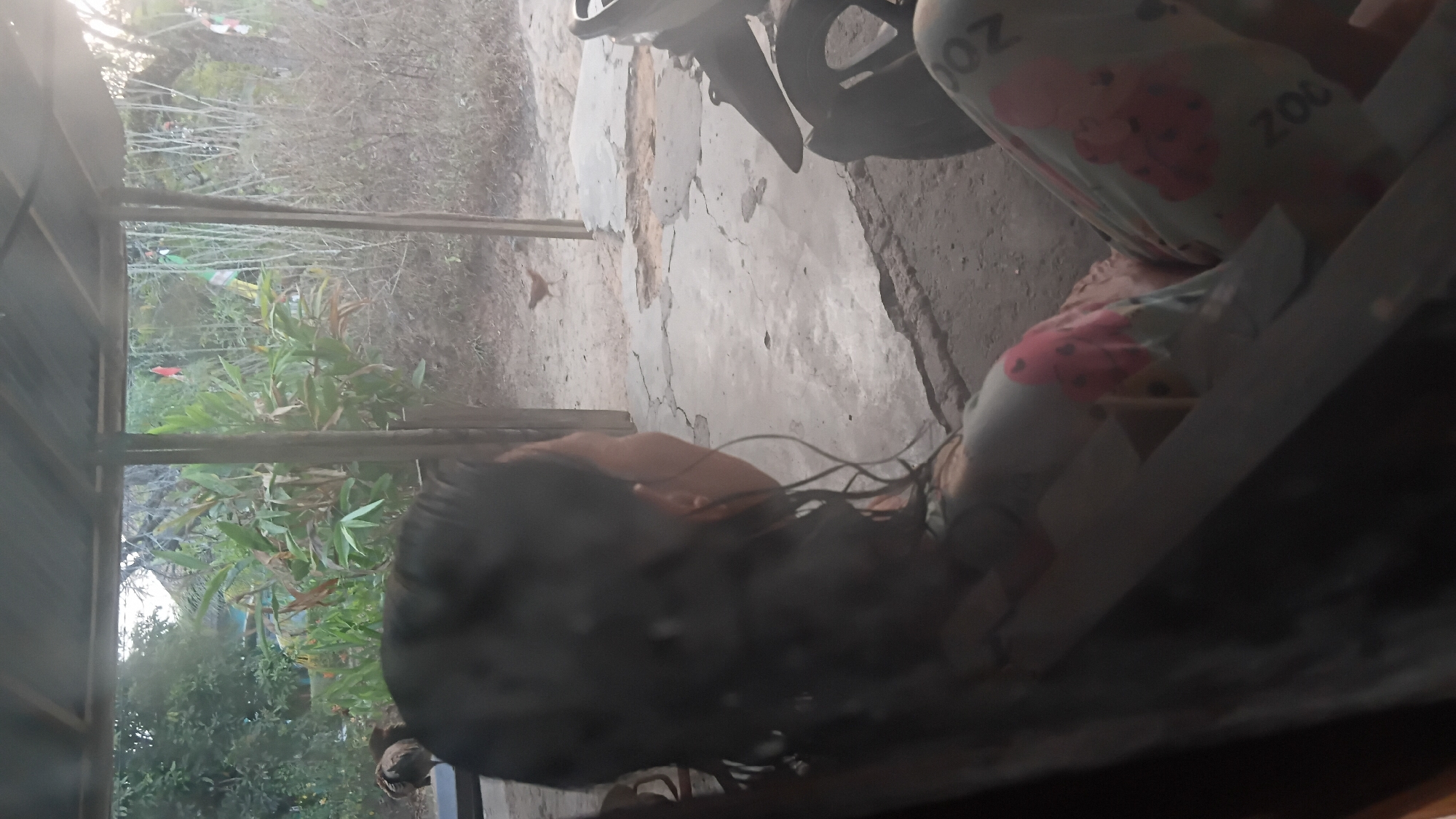 A person is sitting in a chair, viewed from behind and from the side, looking out towards a rural area. The person has dark, long, wet hair and is wearing patterned pants with cartoon bears and the word "ZOO" printed on them. In the background, there are trees, vegetation, and a dirt ground with cracked concrete. A chicken is visible walking on the ground. A motorcycle is partially visible on the right side of the image. The scene appears to be outdoors, possibly on a porch or covered area, with a corrugated metal roof supported by wooden posts. The lighting suggests it is daytime, possibly late afternoon or early morning due to the warm tones and shadows. The location is identified as Prabumulih, Indonesia.