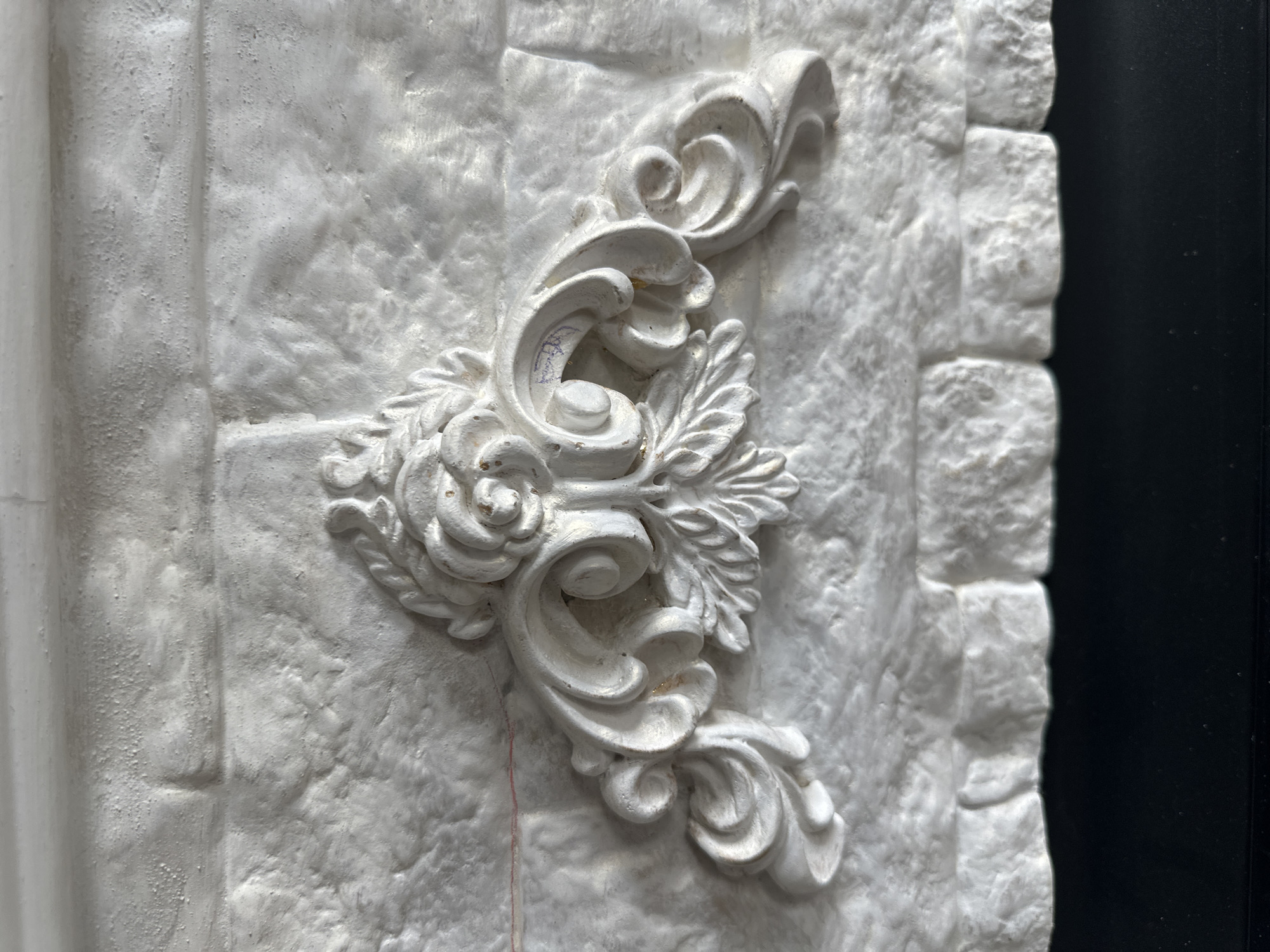 A white, ornate decorative element, resembling a floral acanthus leaf design with swirling scrolls, is affixed to a textured white brick wall. The element is centered and features a prominent rose-like bloom at its apex, with elaborate carvings extending downwards and outwards in symmetrical patterns. The brick wall itself appears to be made of large, irregular blocks with a rough, matte finish, creating a rustic aesthetic. Below the brick wall is a dark, solid surface, possibly a countertop or base. The lighting creates subtle shadows that highlight the three-dimensional qualities of the decorative piece and the brickwork. There are no people or animals present in the image. The scene is static and appears to be an interior architectural detail.