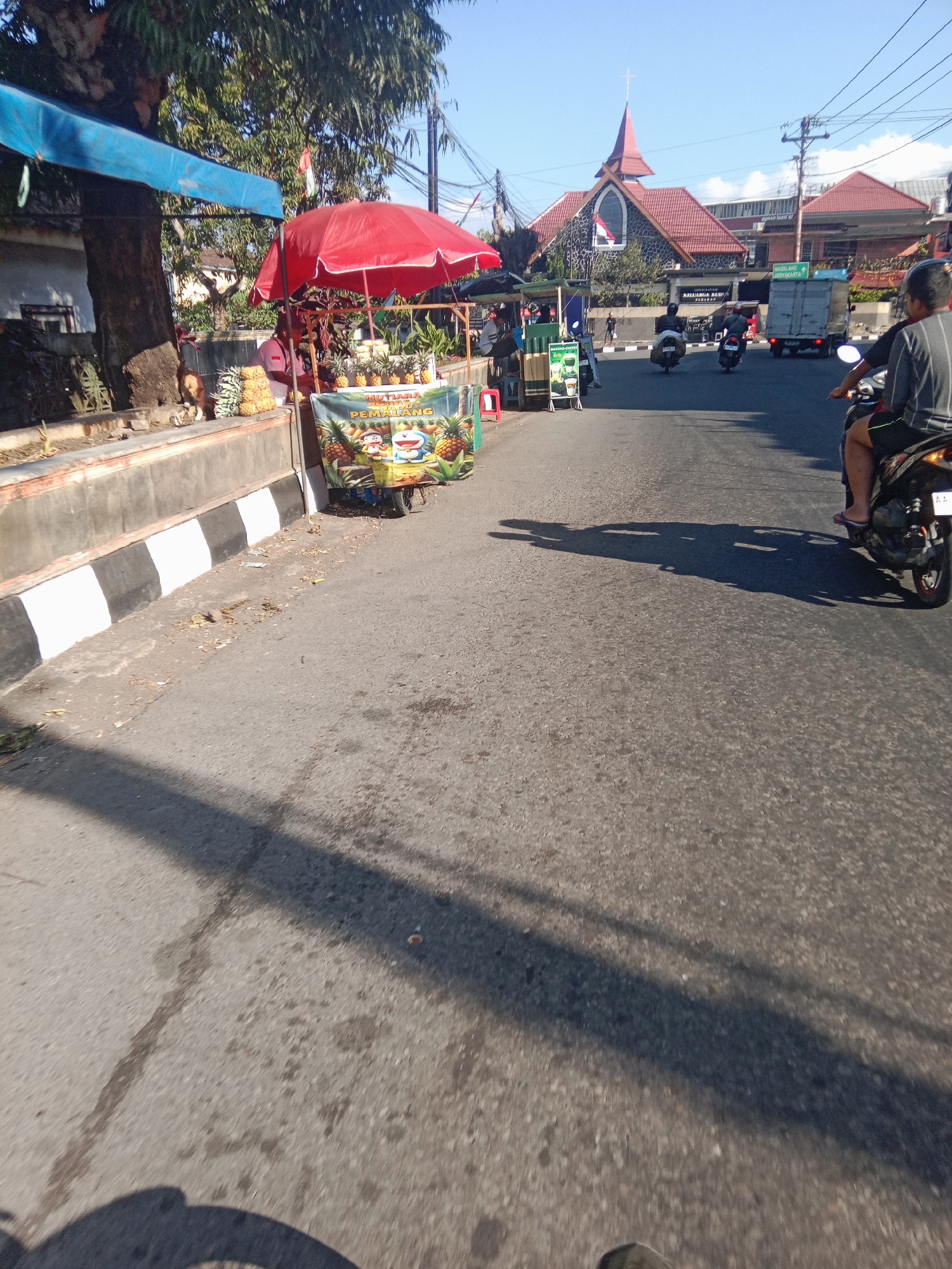 This is a street-level view in Parakan, Indonesia, on a sunny day. The scene is dominated by a road with a visible shadow from a moving object, likely a motorcycle, in the foreground. To the left, a sidewalk area is bordered by a concrete barrier with black and white striped markings. Behind this barrier, a large tree stands with a blue awning extending over it. Adjacent to the tree, a fruit stand is set up under a red umbrella, displaying pineapples and other produce. A banner on the stand features cartoon characters and text, possibly advertising the fruit. A person in a red shirt is visible behind the fruit stand. Further down the street, a church with a distinctive red roof and steeple is prominent. Other buildings and a truck are also visible, suggesting a commercial or residential area. Several motorcycles are on the road, with two riders clearly visible. The overall impression is a typical street scene in an Indonesian town, with local commerce and daily life unfolding.