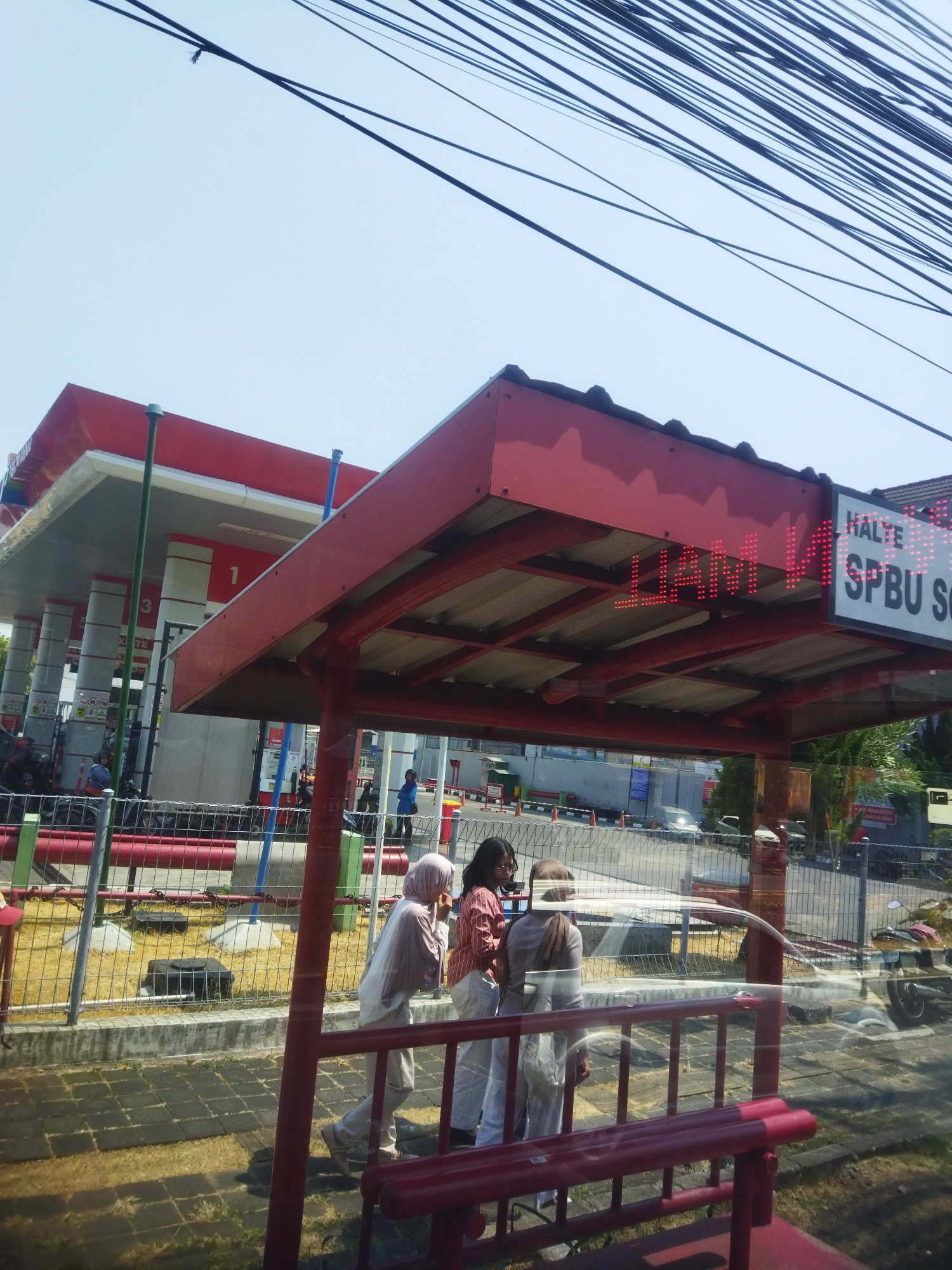 A street-level view captures a red bus stop shelter with a digital display showing "HALTE SPBU SQ". The display also shows "LIAM LI". Three women are standing under the shelter, two of them wearing hijabs. The woman on the left is wearing a light brown hijab and a light beige outfit. The woman in the middle is wearing a brown hijab and a red and white striped shirt with white pants. The woman on the right is wearing a brown hijab and a white outfit. In the background, a red and white gas station with multiple pumps is visible. There are several motorcycles parked in front of the gas station. The ground is paved with gray tiles, and there is a yellow sandy area with some dark rectangular objects behind a metal fence. Overhead, a network of power lines is present against a pale blue sky. The scene appears to be on a sunny day. This location is identified as Grogol, Indonesia, specifically a bus stop at a gas station.