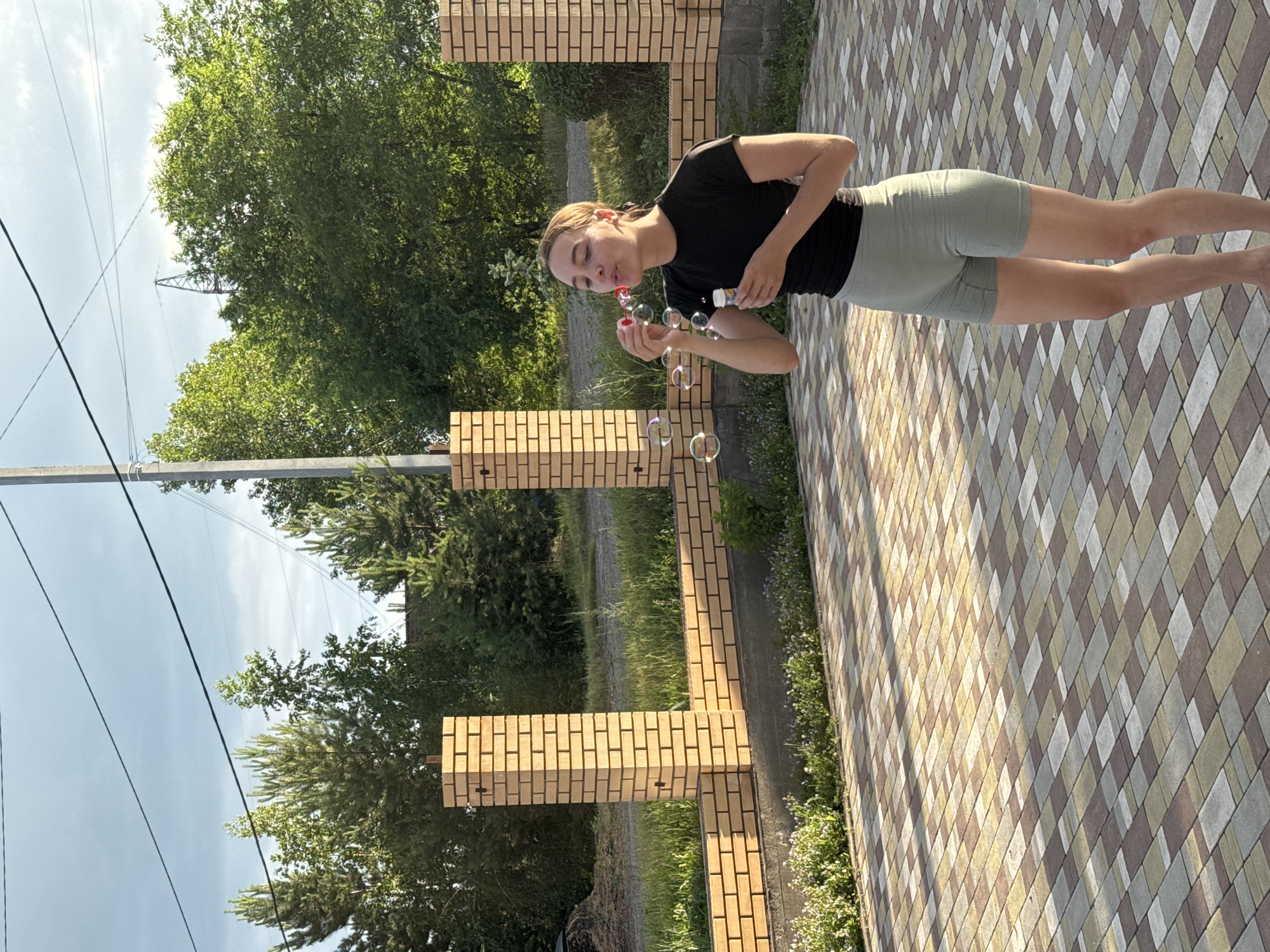 A young woman is blowing bubbles in a backyard setting. She is wearing a black t-shirt and light green shorts. She is holding a bottle of bubble solution and a wand, from which several bubbles are emerging. The woman has her eyes closed and is focused on blowing the bubbles. The background shows a tiled patio, brick pillars, and trees. The scene appears to be outdoors during the daytime, with bright sunlight casting shadows on the patio. The overall mood is playful and relaxed. There are power lines visible in the upper left portion of the image, and what appears to be a construction crane in the distance behind some trees. The location context is Minusinsk, Russia.